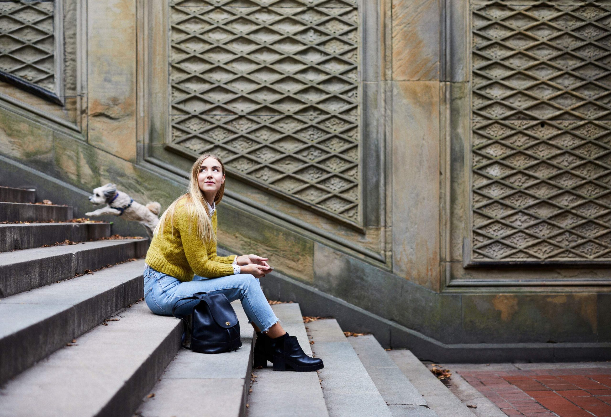 A woman sits on stone steps with a running dog behind her.