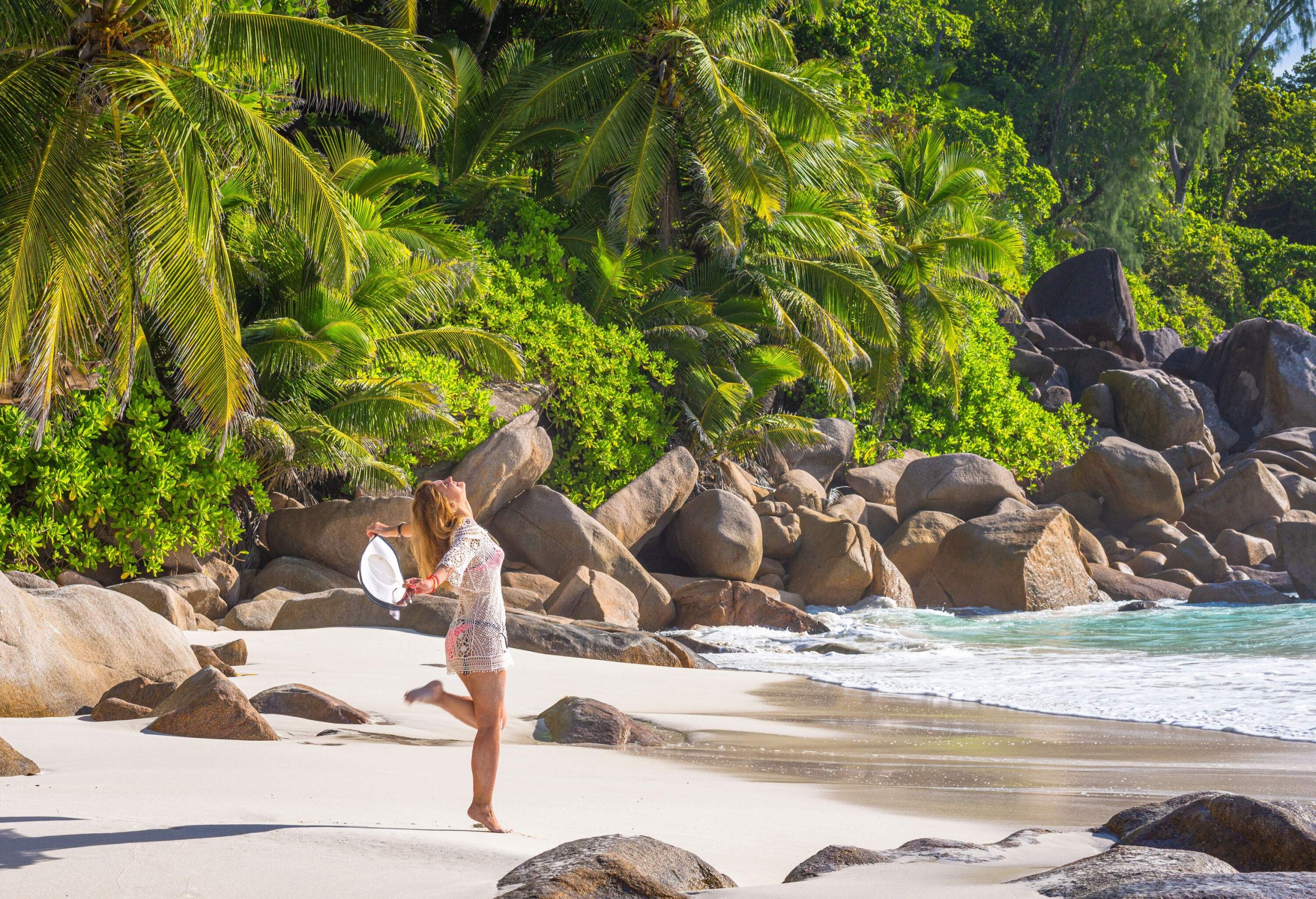 A cheerful woman spreads her arms as she stands on a beach with big boulders and lush tropical foliage.