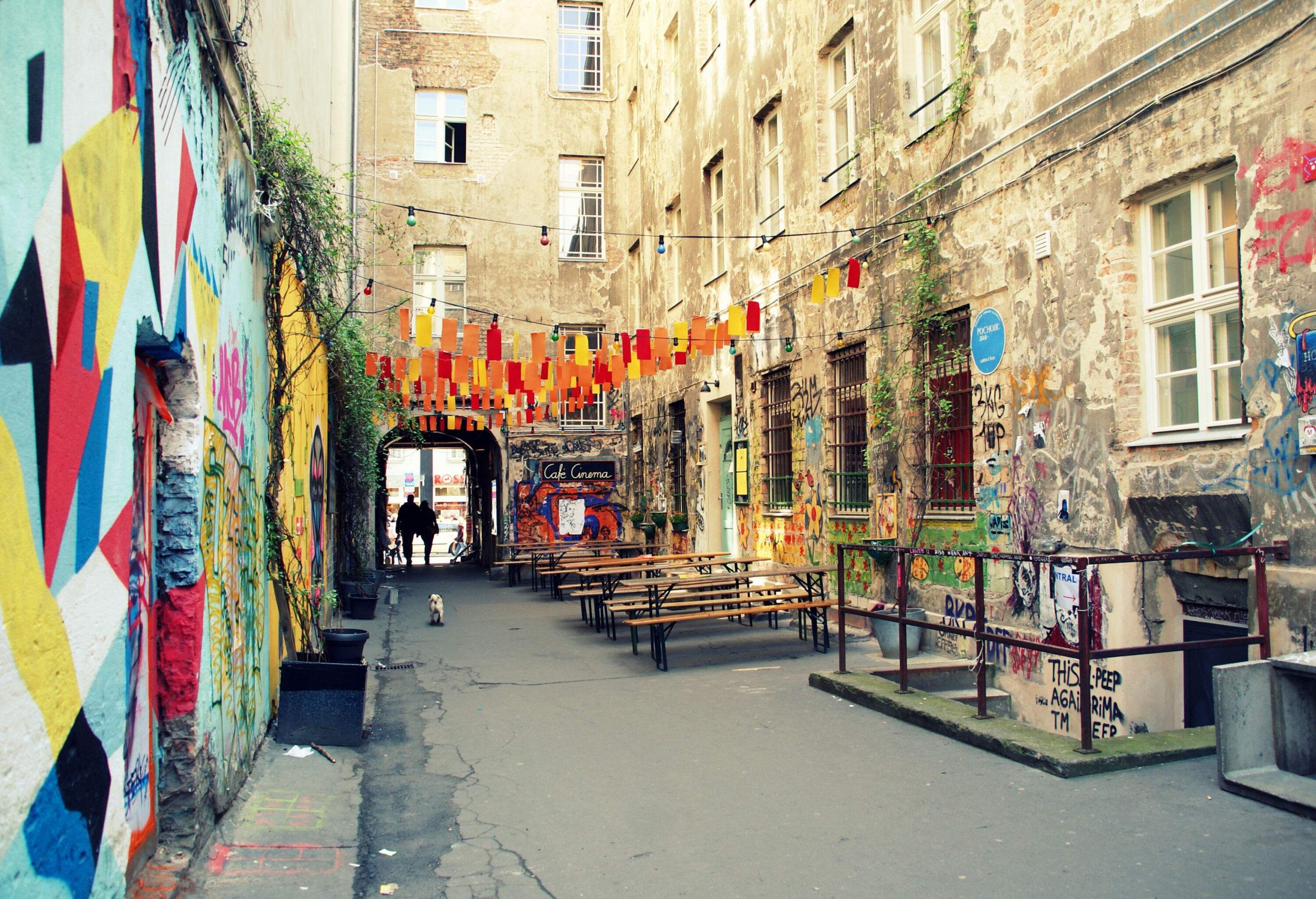 A narrow alley with outdoor picnic tables under hanging festival banners between tall and old buildings.