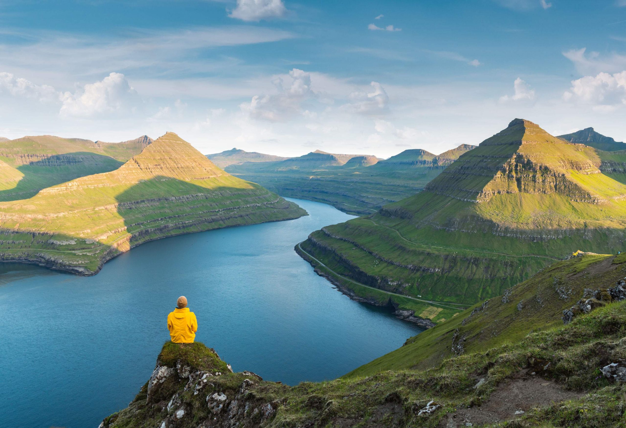 A person in a yellow jacket sits on a narrow cliff overlooking the calm waters below the crags of the island.
