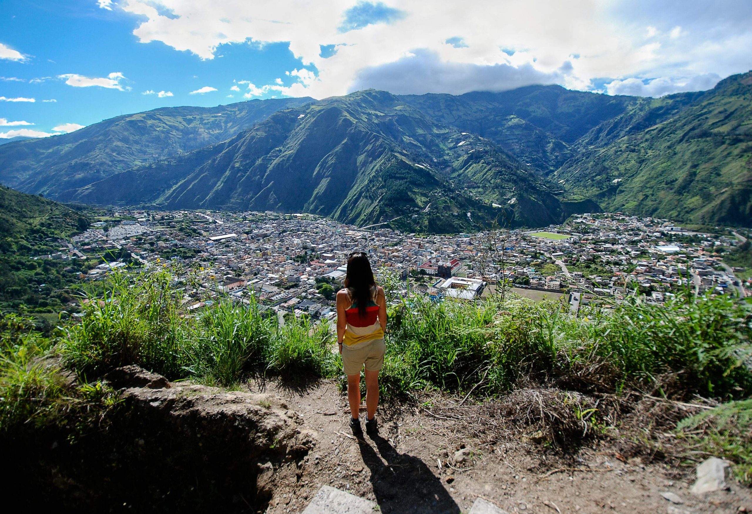 A woman standing on a sandy slope with tall grass, looking over the structures spread across the valley.