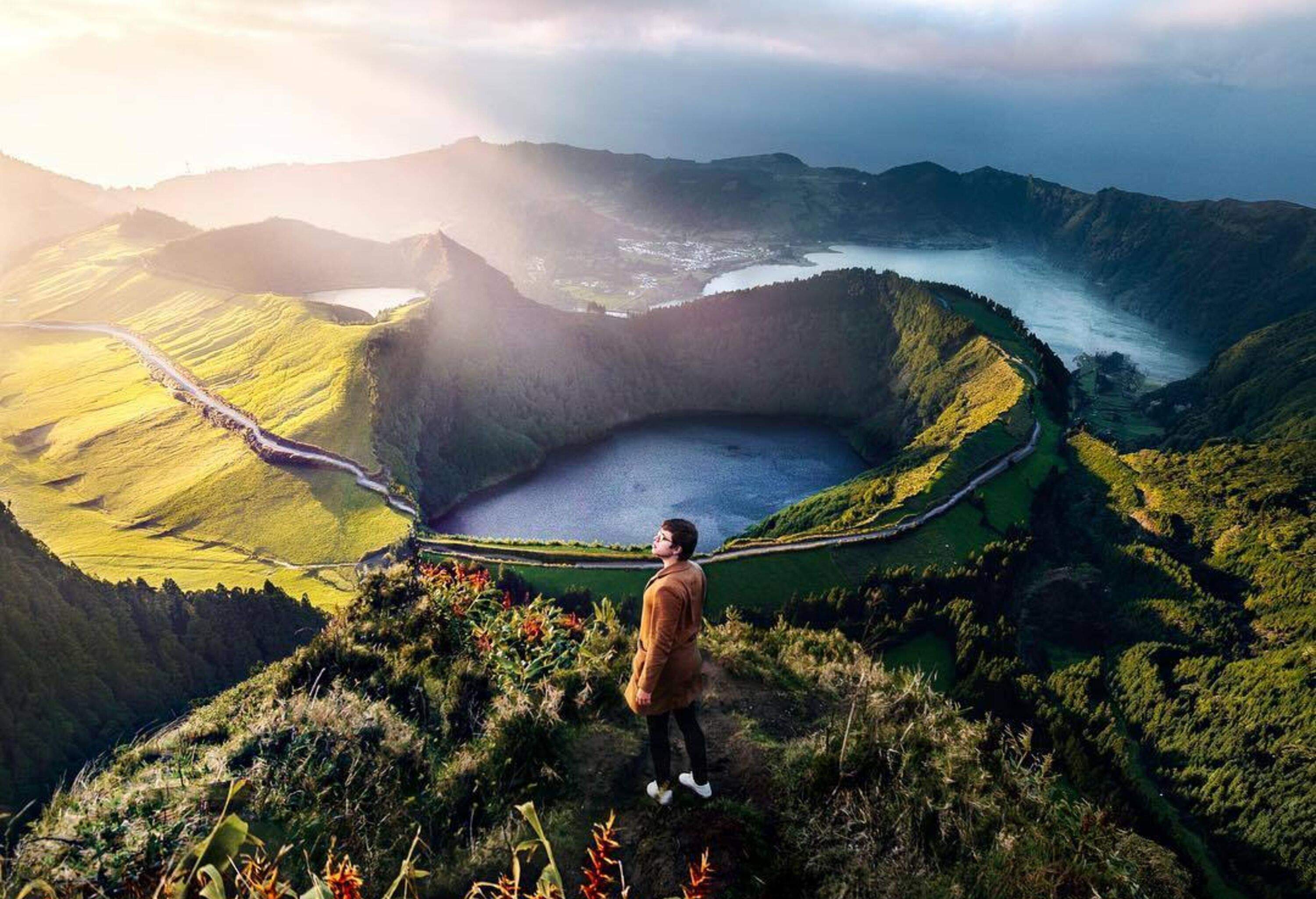 A male tourist stands tall on top of a lush mountain overlooking the spectacular crater lakes against the scenic sky.