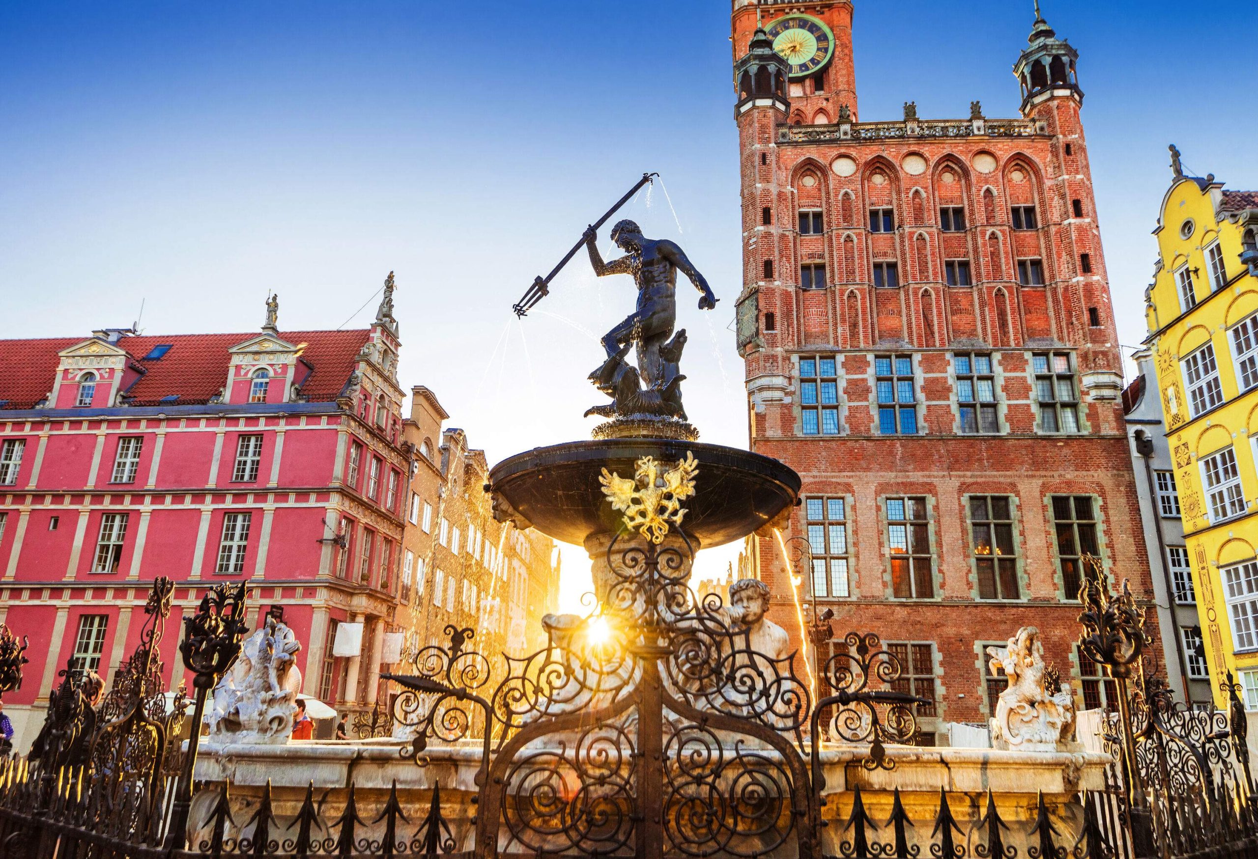 A beautiful fountain with statues in the centre of a city square surrounded by colourful and classical buildings.