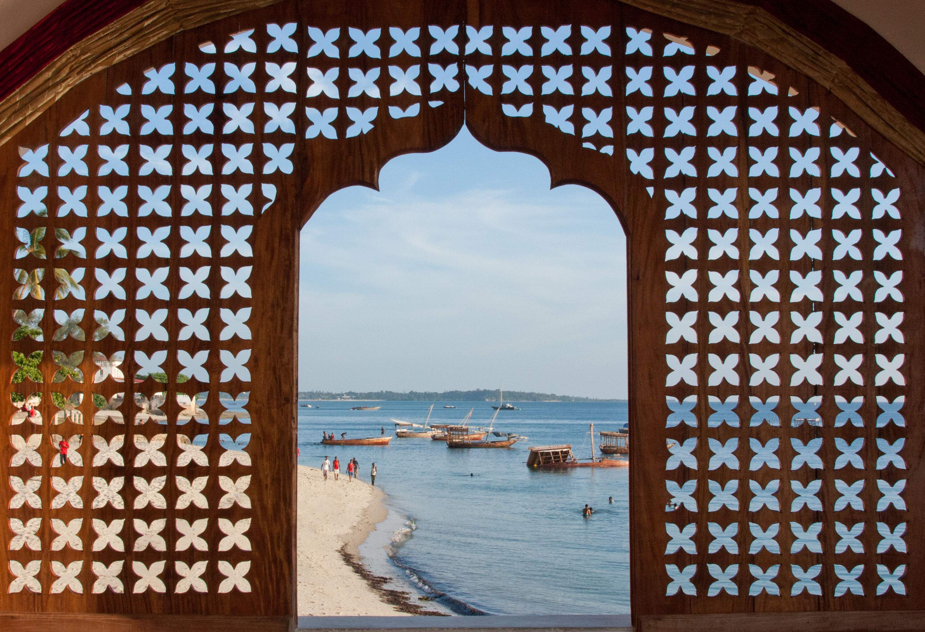 A tranquil beach lined with moored boats, viewed through a latticework window of a hotel.