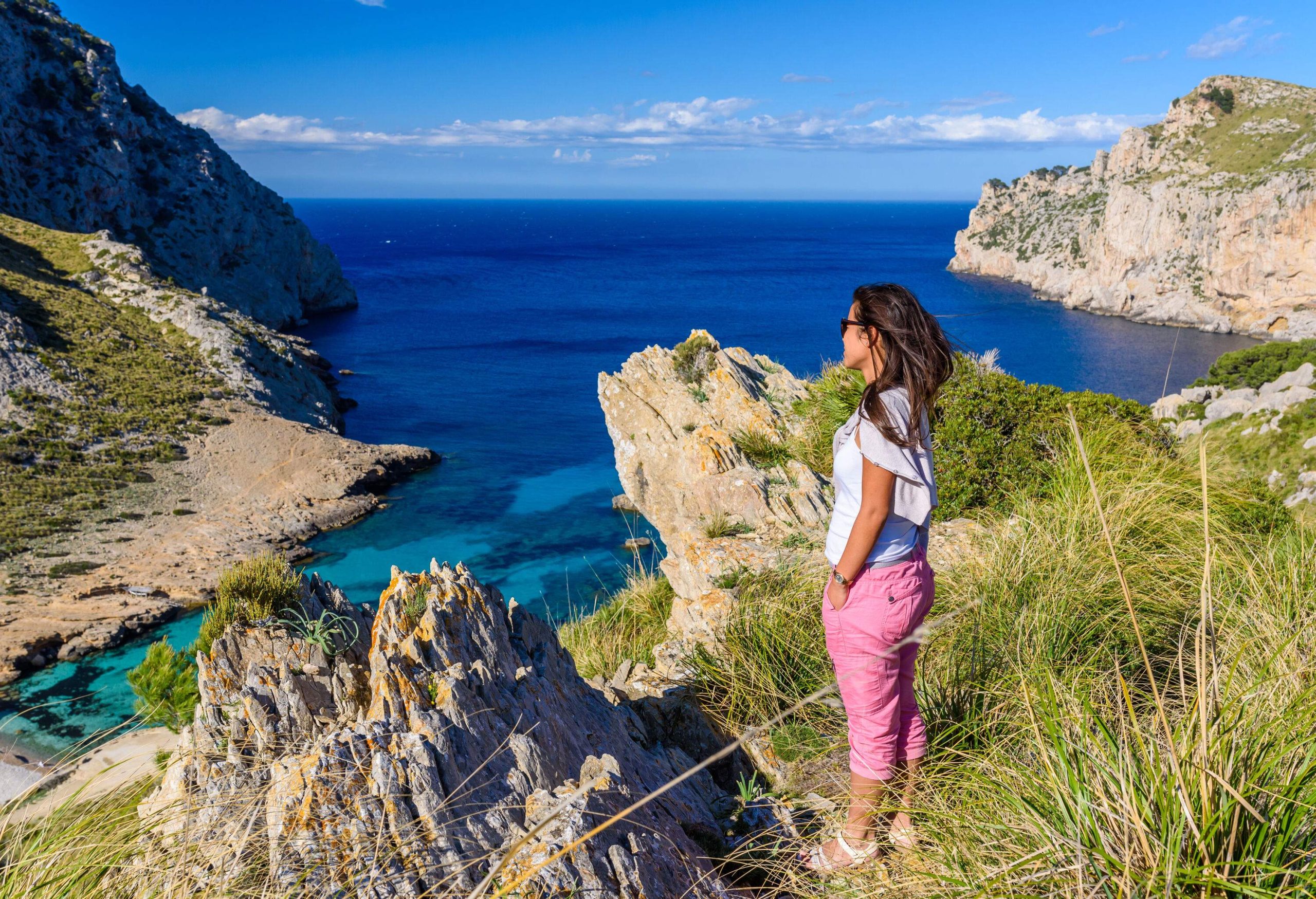 A person standing on a cliff looking out over the blue sea and rocky cliffs.