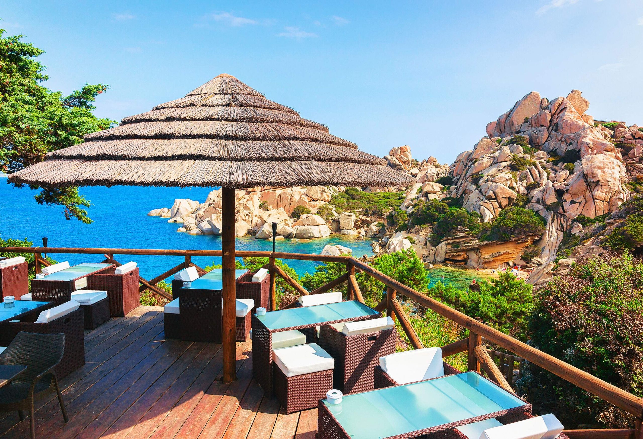 An outdoor deck overlooking the beach with wicker tables and chairs shaded by a straw beach umbrella.