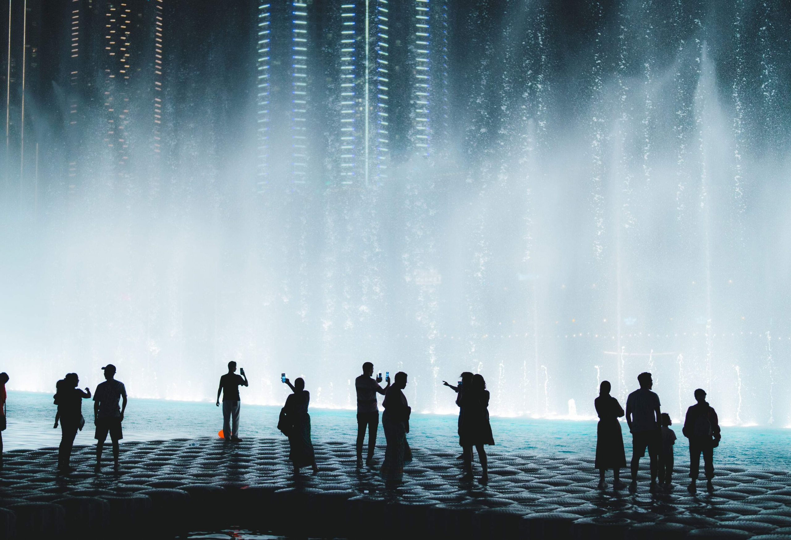 A captivating silhouette of people, their gazes fixed upon the mesmerising fountain performance under the night sky.