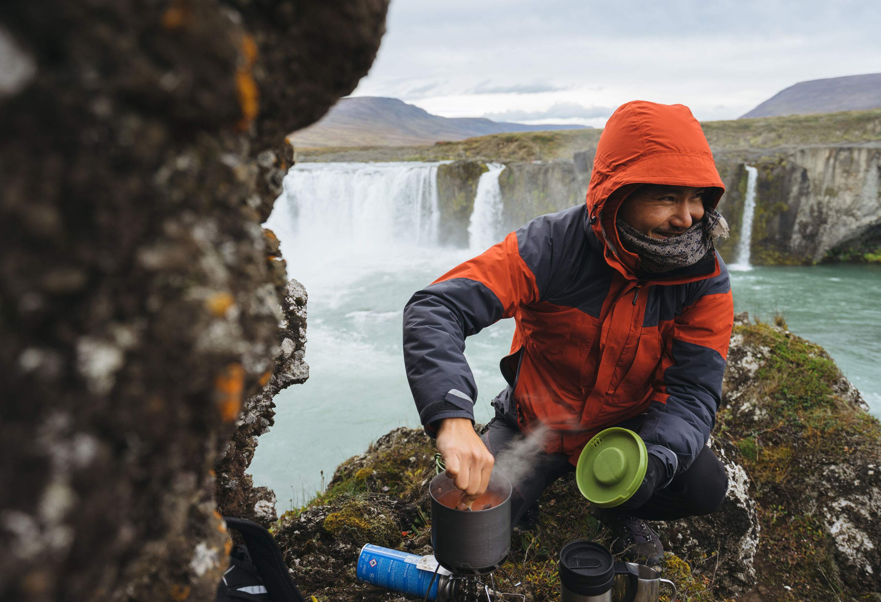 A male camper cooking on a camping stove with a beautiful waterfall behind him.