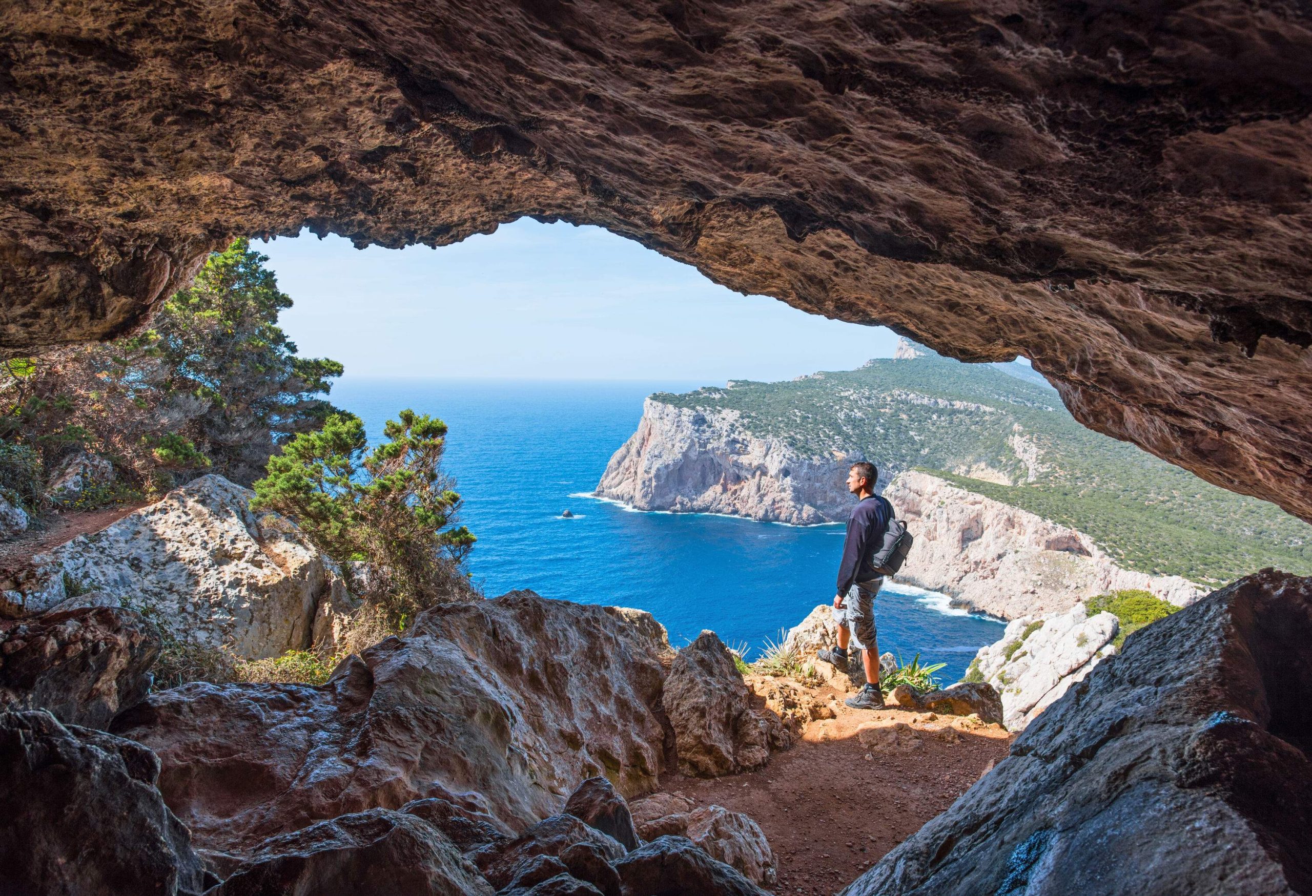 A male hiker standing along the edge outside of a cave overlooking the blue sea.