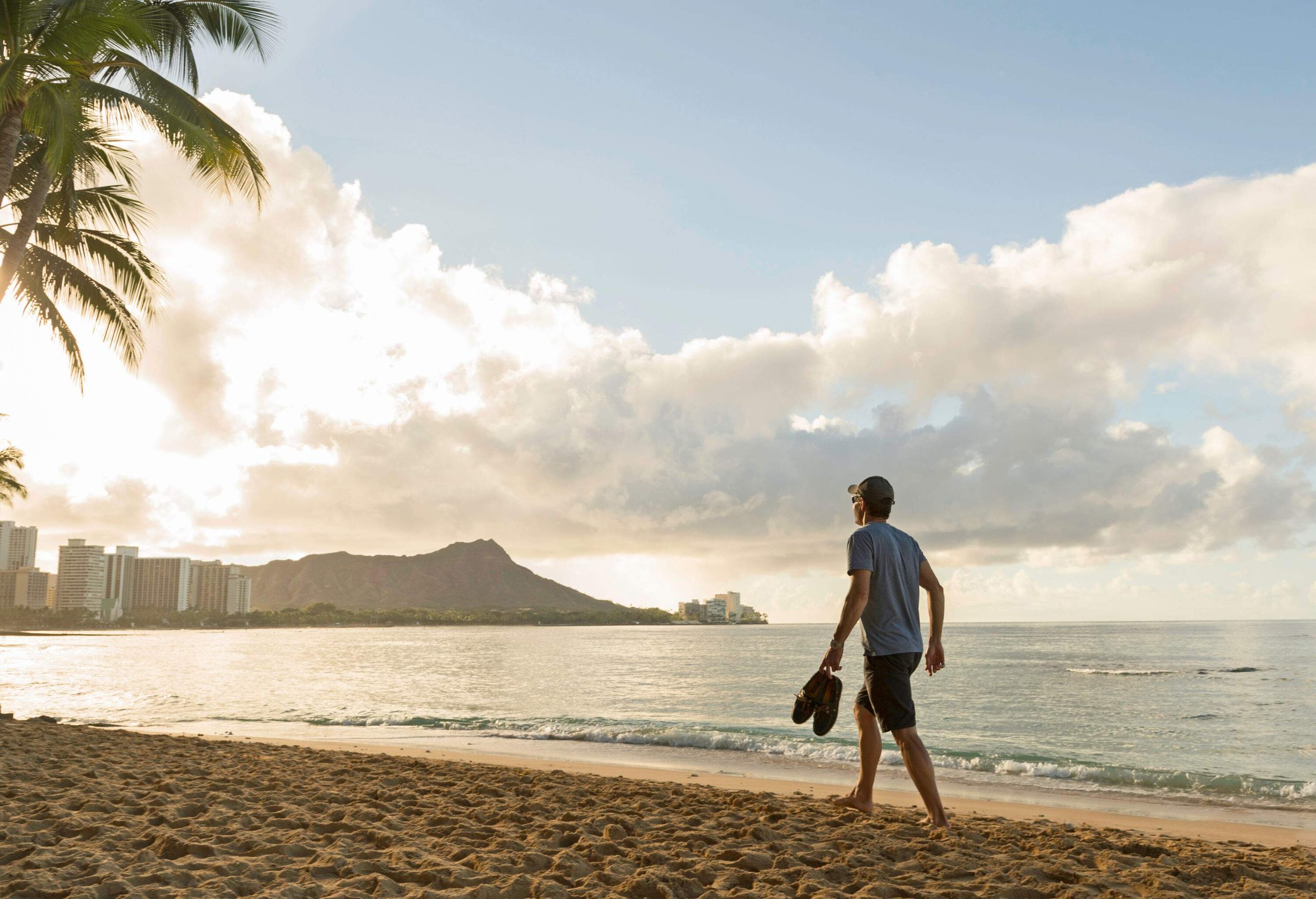 A man strolls down a beach while carrying a pair of shoes.