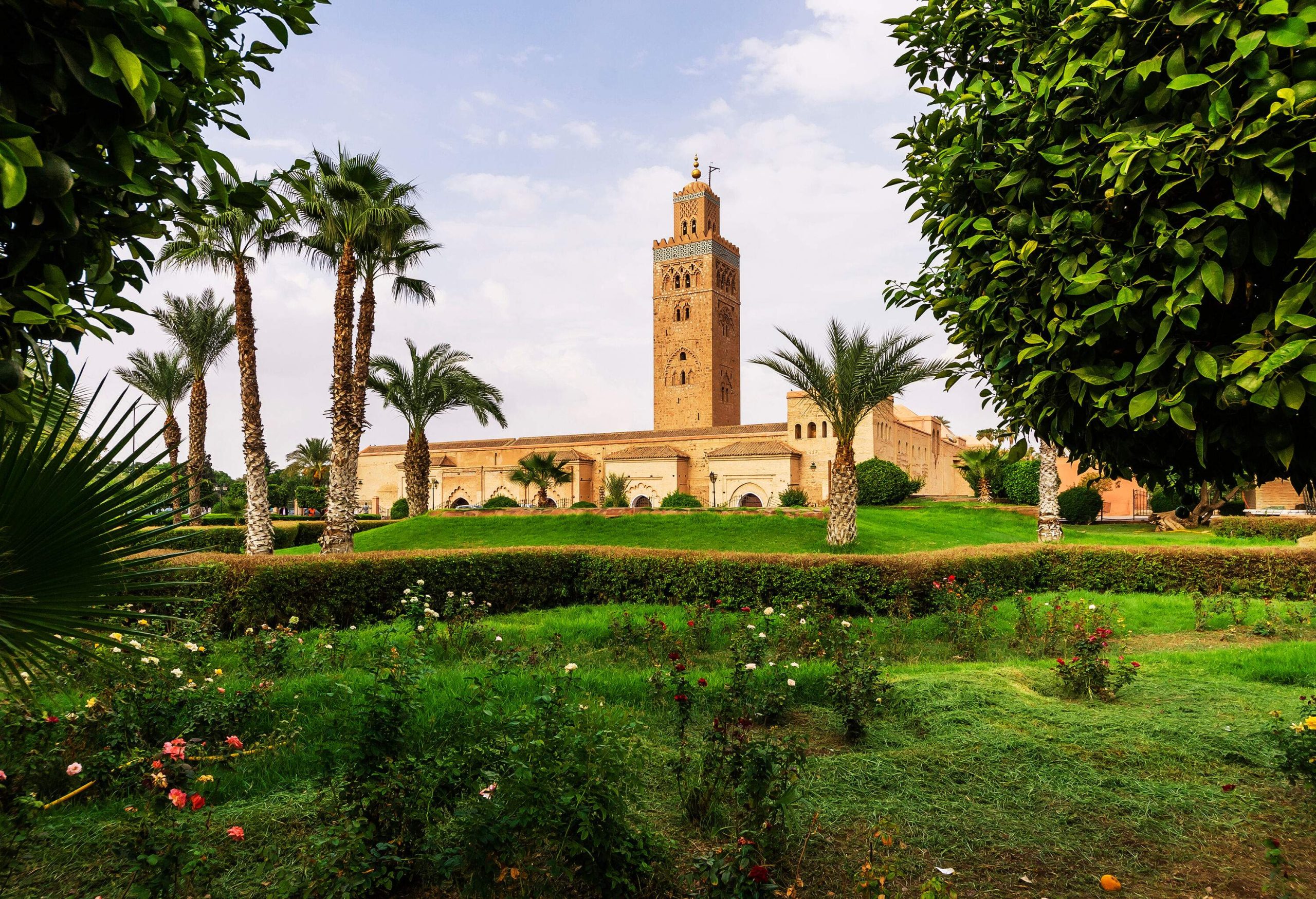 A mosque with its protruding minaret tower topped by a spire and metal orbs along a lush green garden.