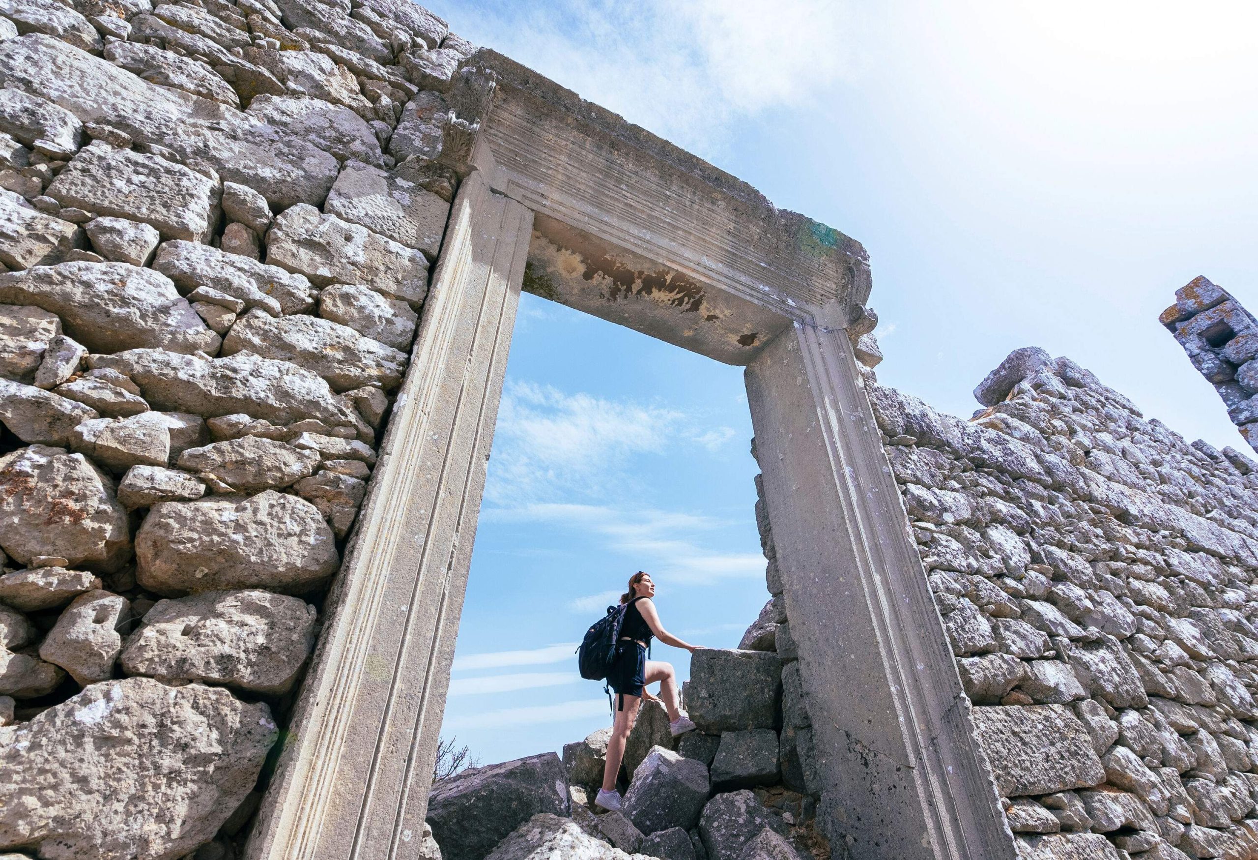 A young woman climbing over large blocks of rocks behind an ancient entry gate.