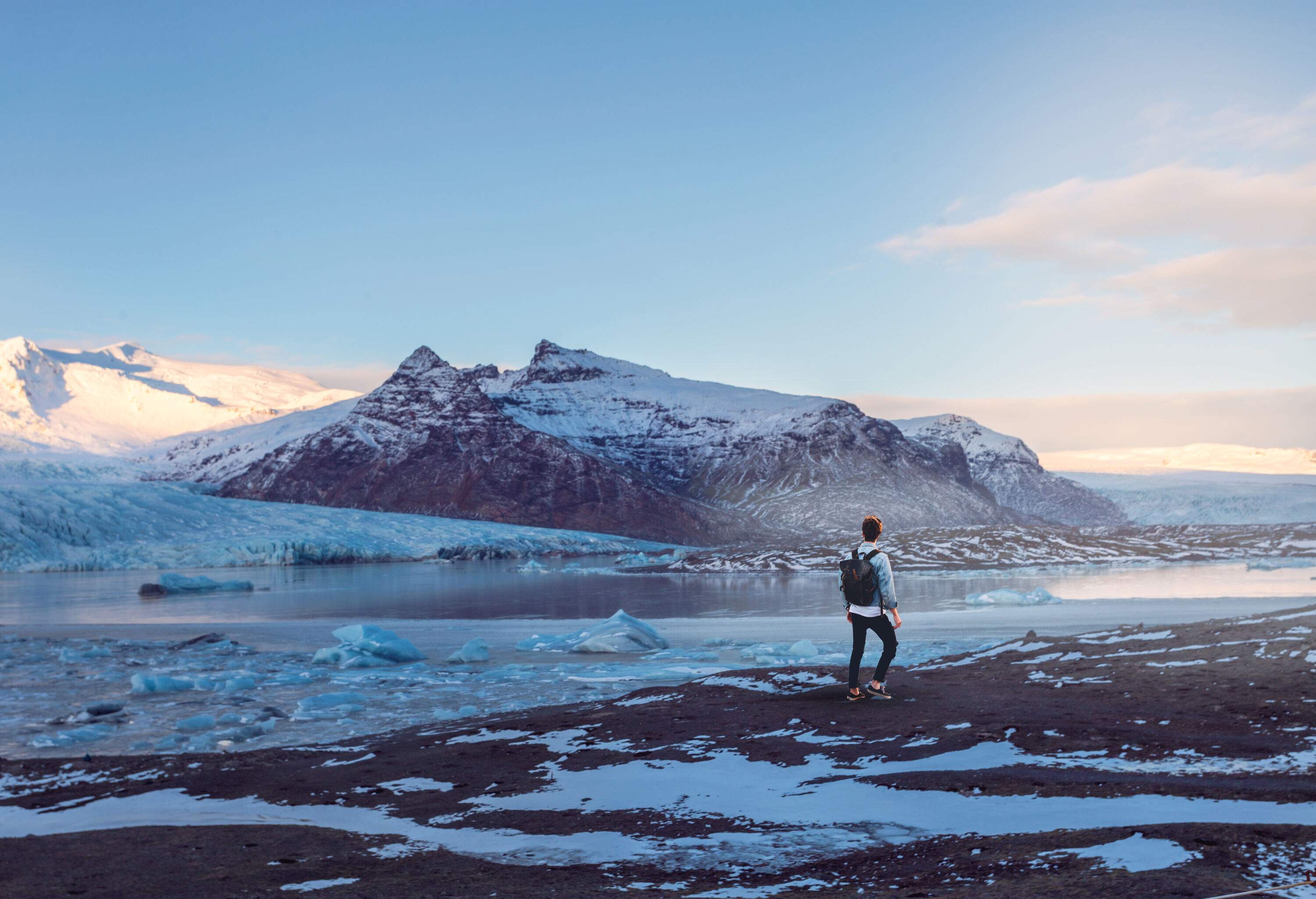 A person walks along the beach of a serene lake surrounded by snowed grounds, with a snow-covered rocky rock formation rising up in the distance.