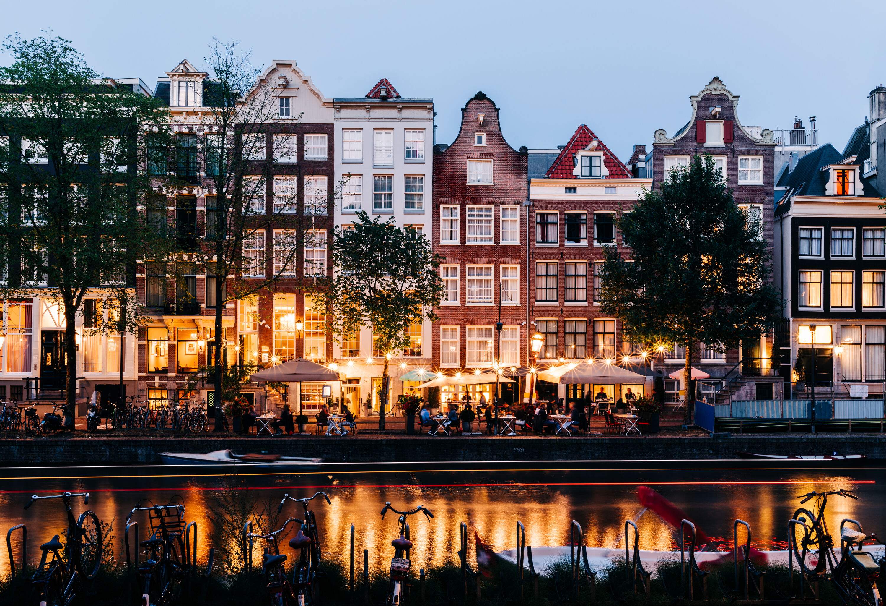 People dine by the river next to traditional tall, narrow, brightly lit Amsterdam canal houses.