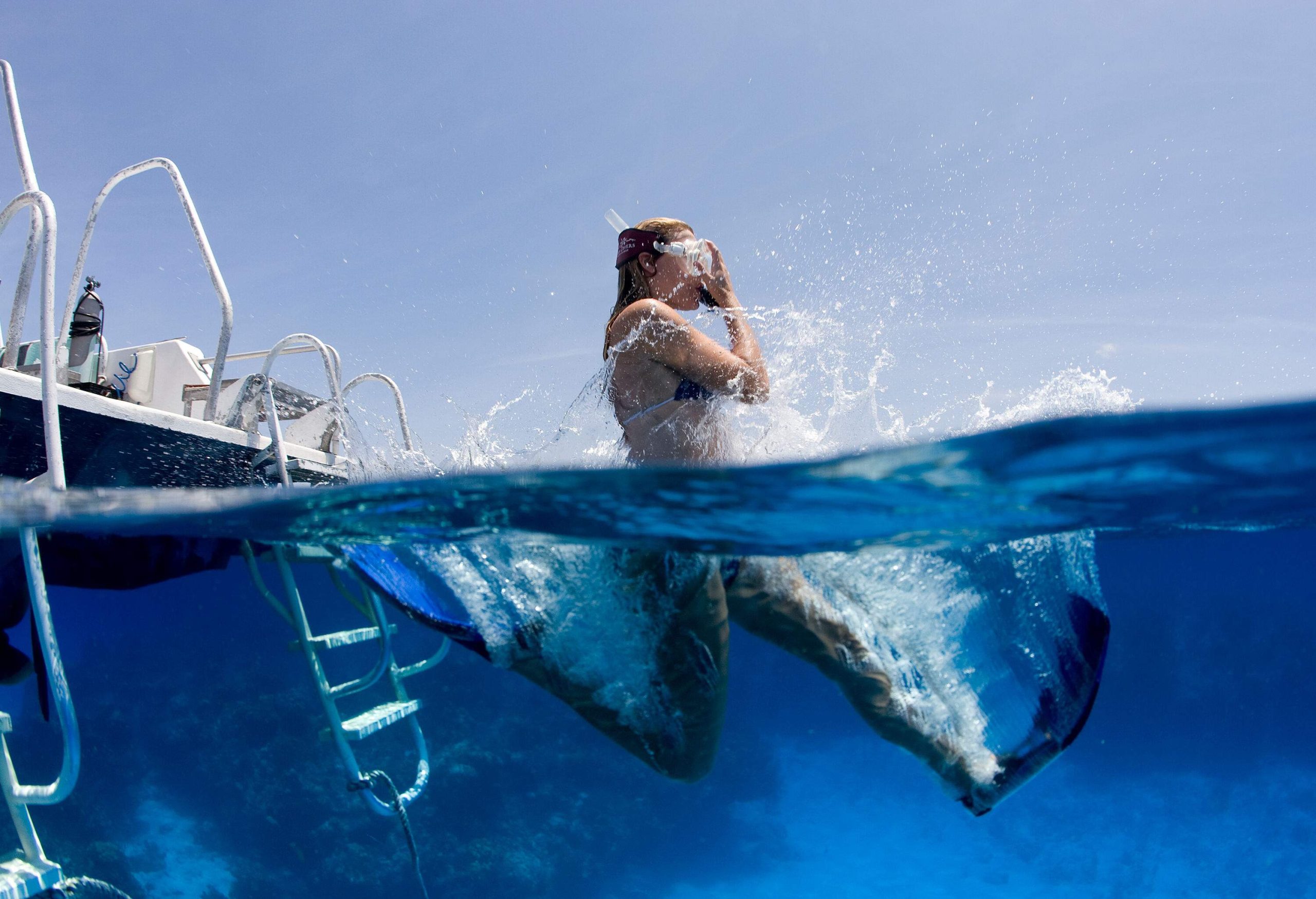 A female wearing snorkelling gear jumps down the blue sea from the boat.