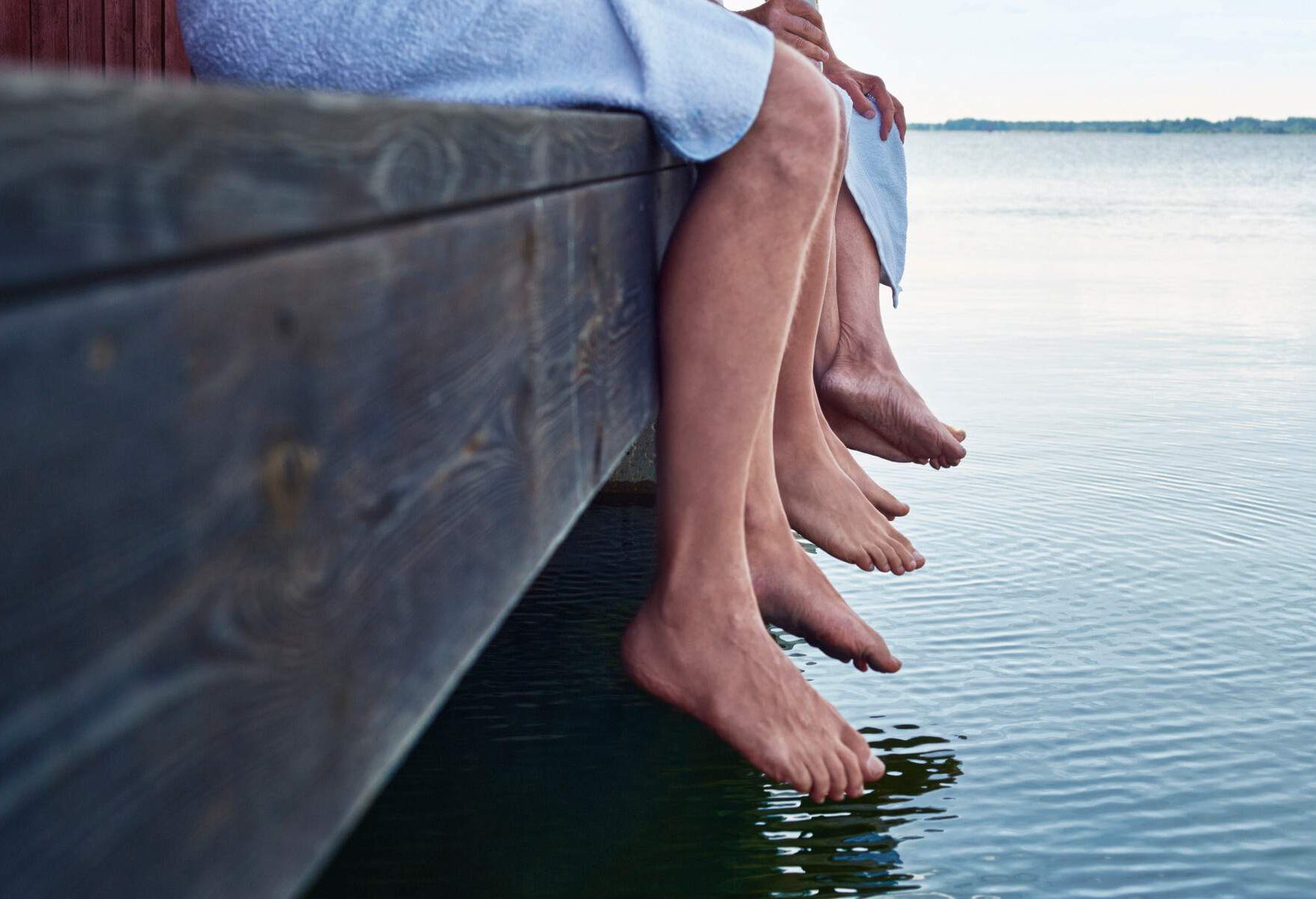 Three pairs of legs hanging on a wooden platform above a lake.