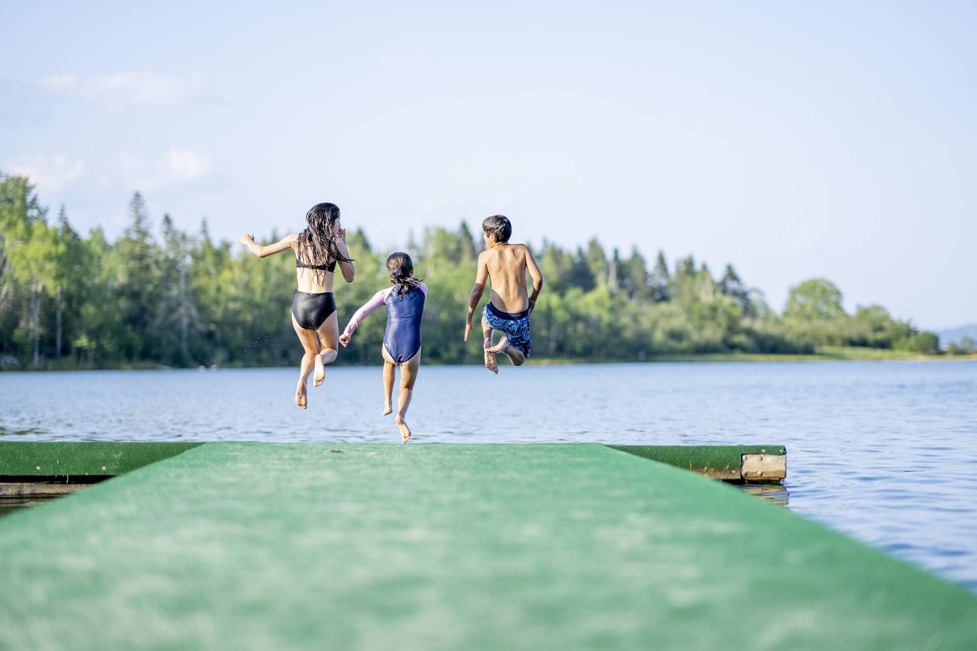 Three children in swimsuits leap into a lake from a jetty.