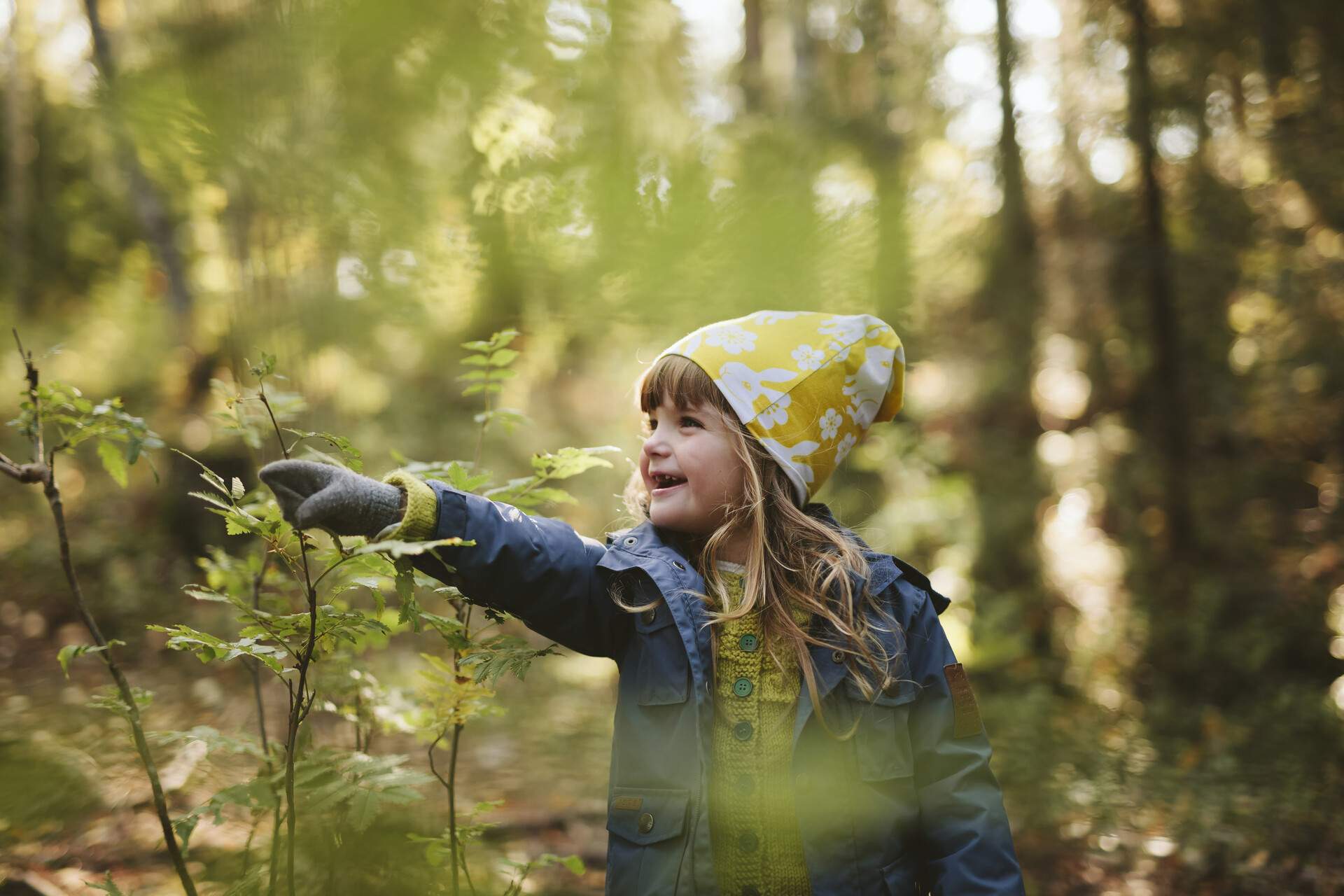 A little girl dressed in warm clothing pointing at a plant during forest hike.

