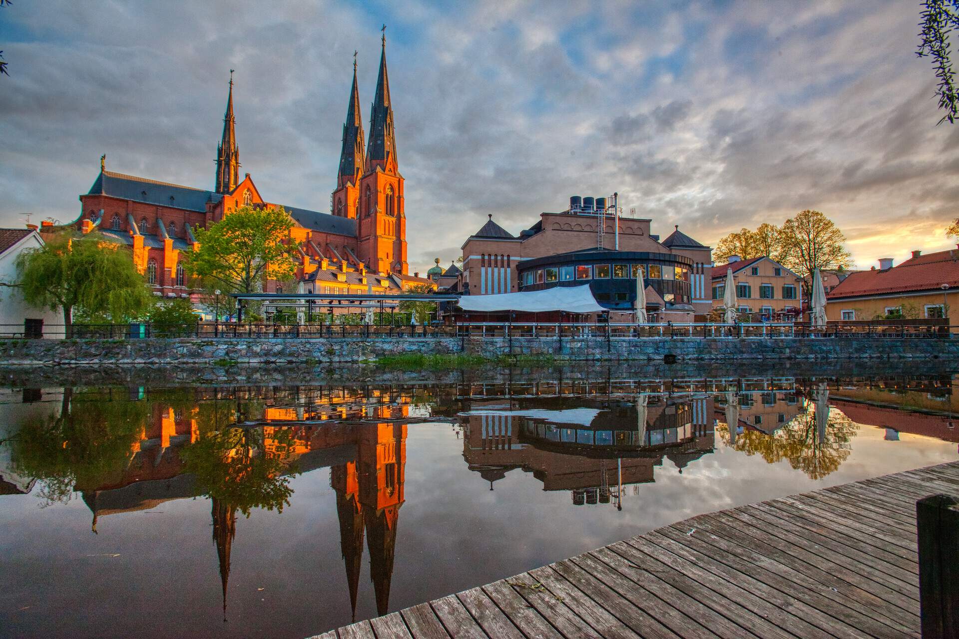 A two-towered church with three spires lies beside a building reflected on the river's surface.
