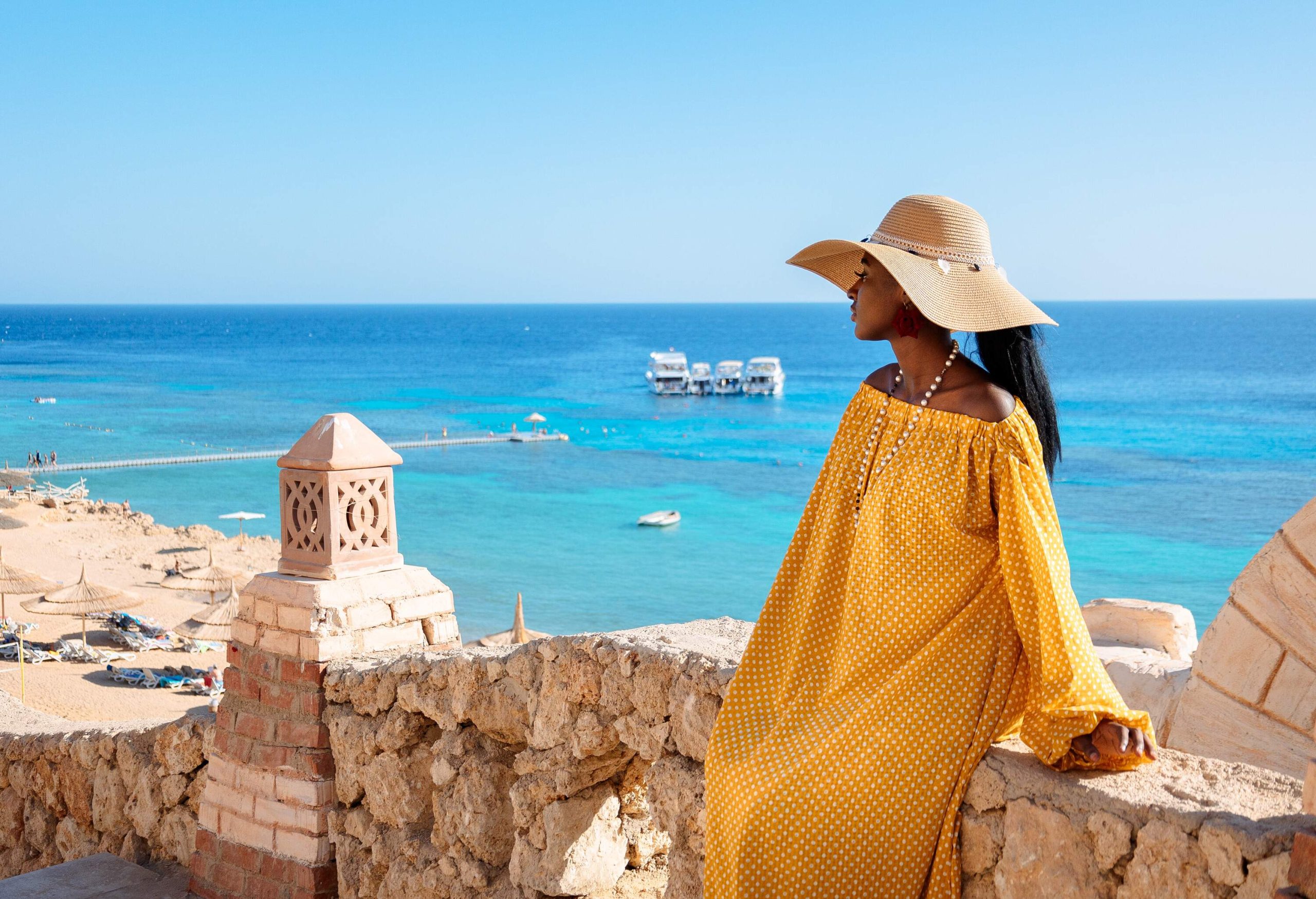 An African lady tourist sits on the stone fence, viewing the protruding dock into the yachts on the turquoise sea.