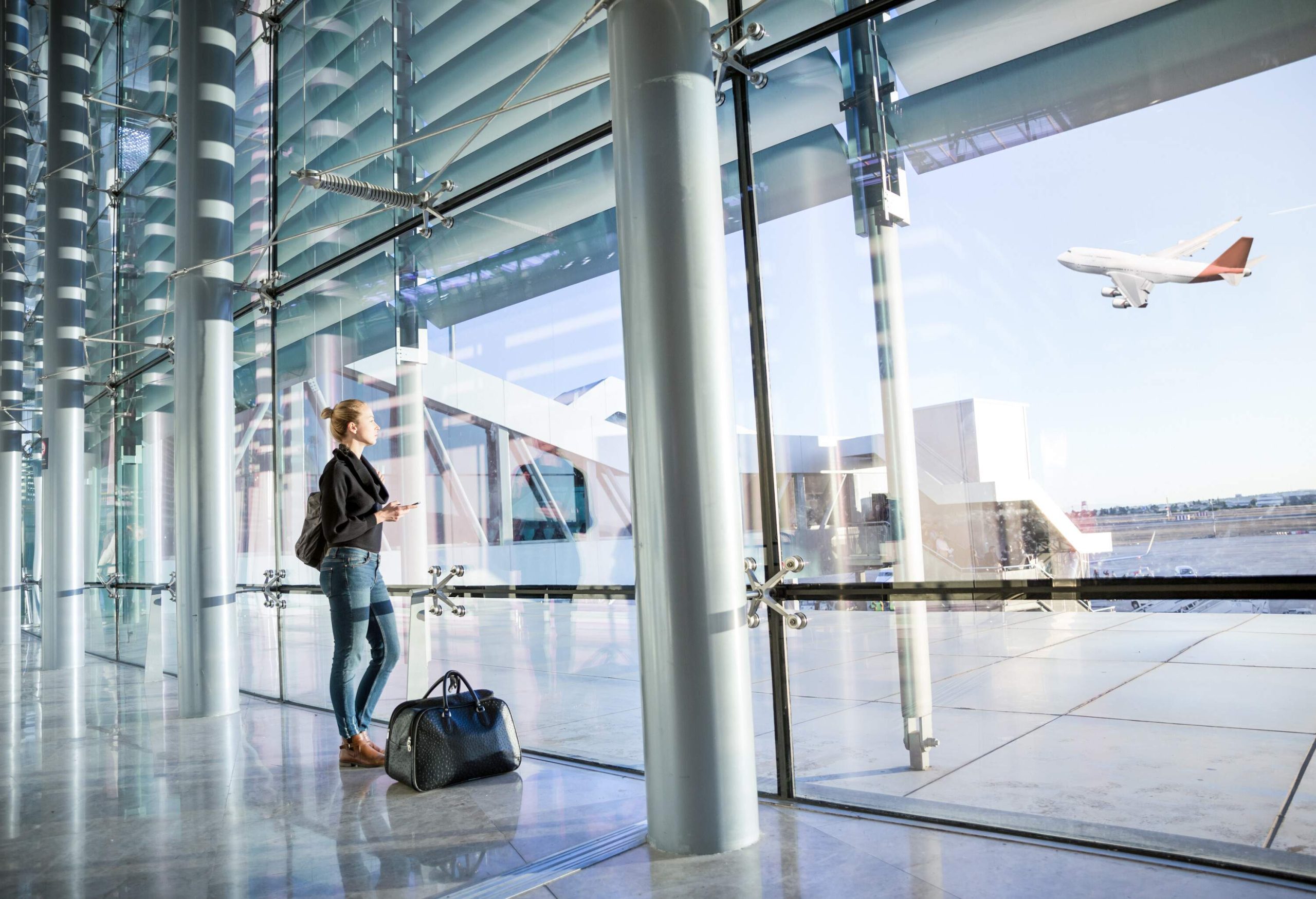 A lady passenger holding her phone stands beside the big glass window and watches the airplane take off.