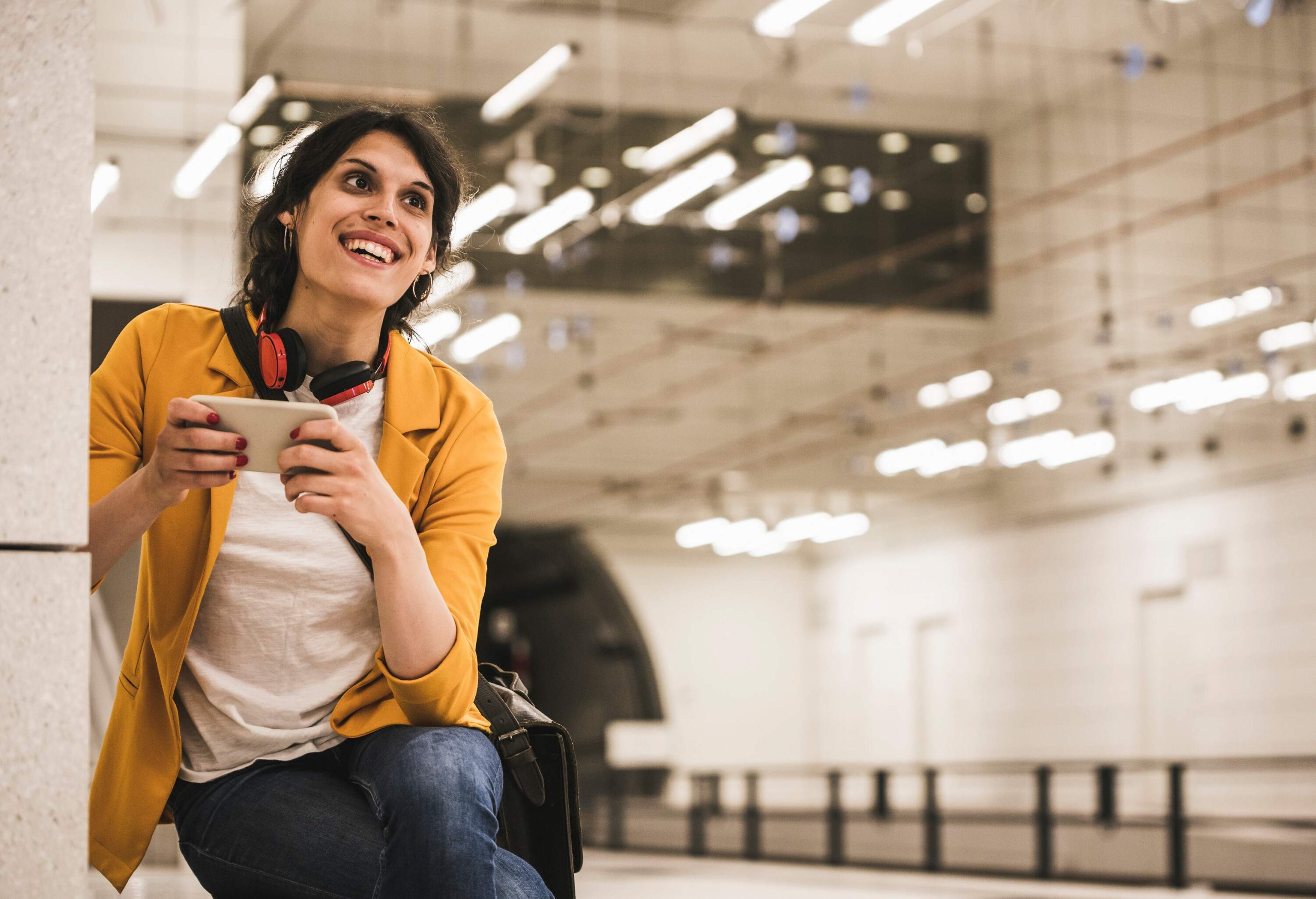 A smiling and confident woman holds a phone and gazes into the distance at a subway station.