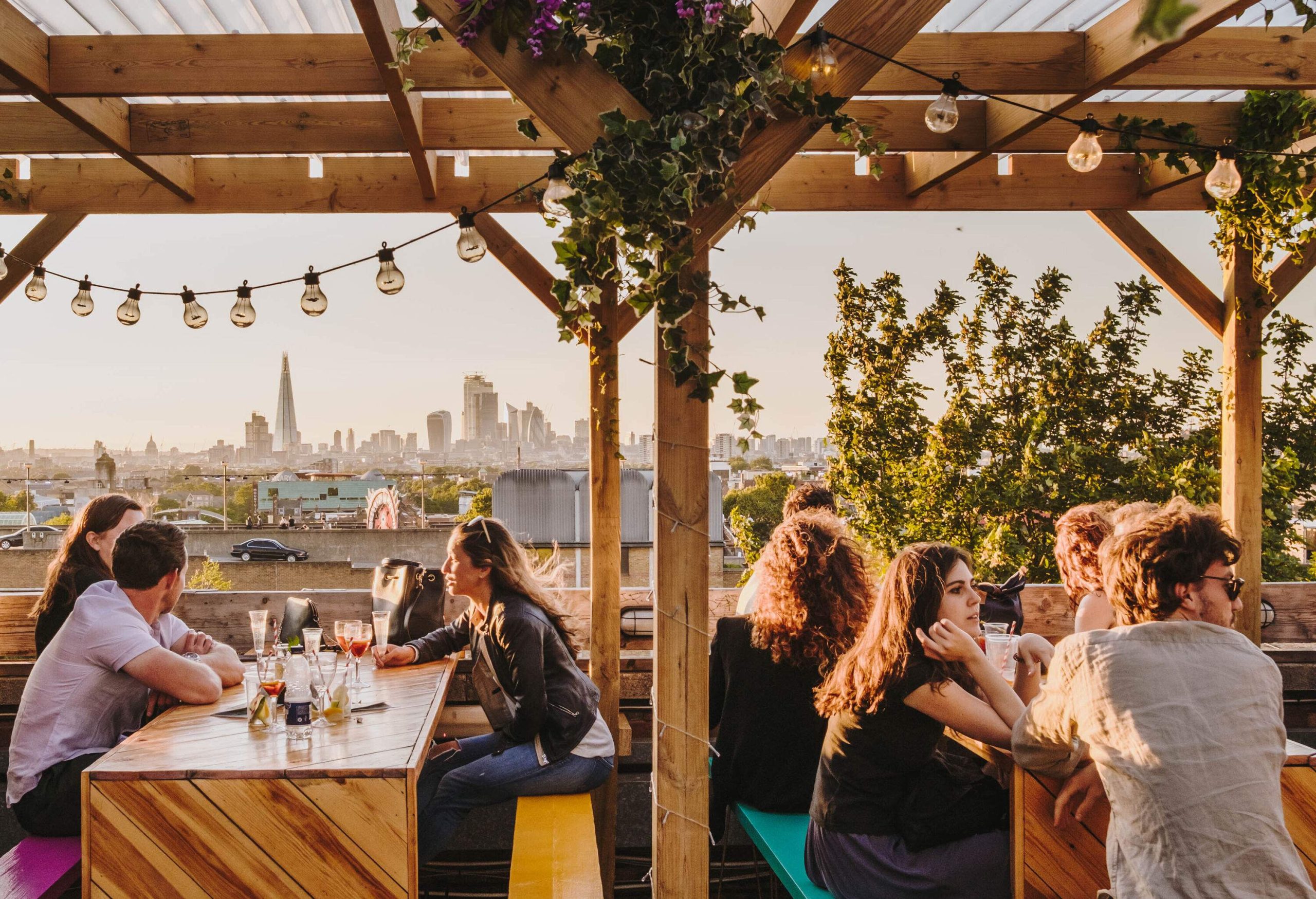 Groups of people hanging out on a rooftop bar decorated with string lights along with providing a view of the city.