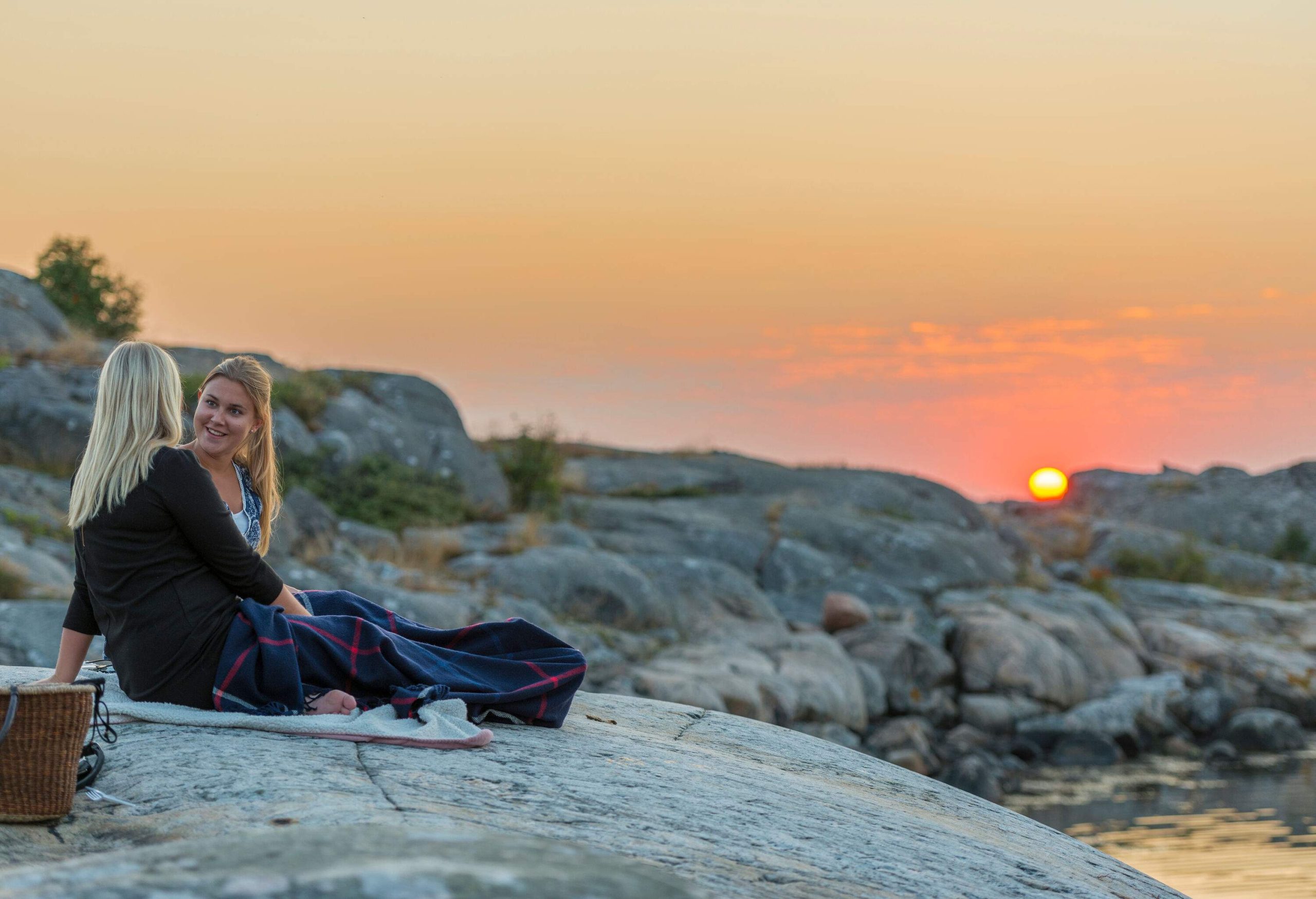 Two women are picnicking on a large boulder against a stone background at sundown.
