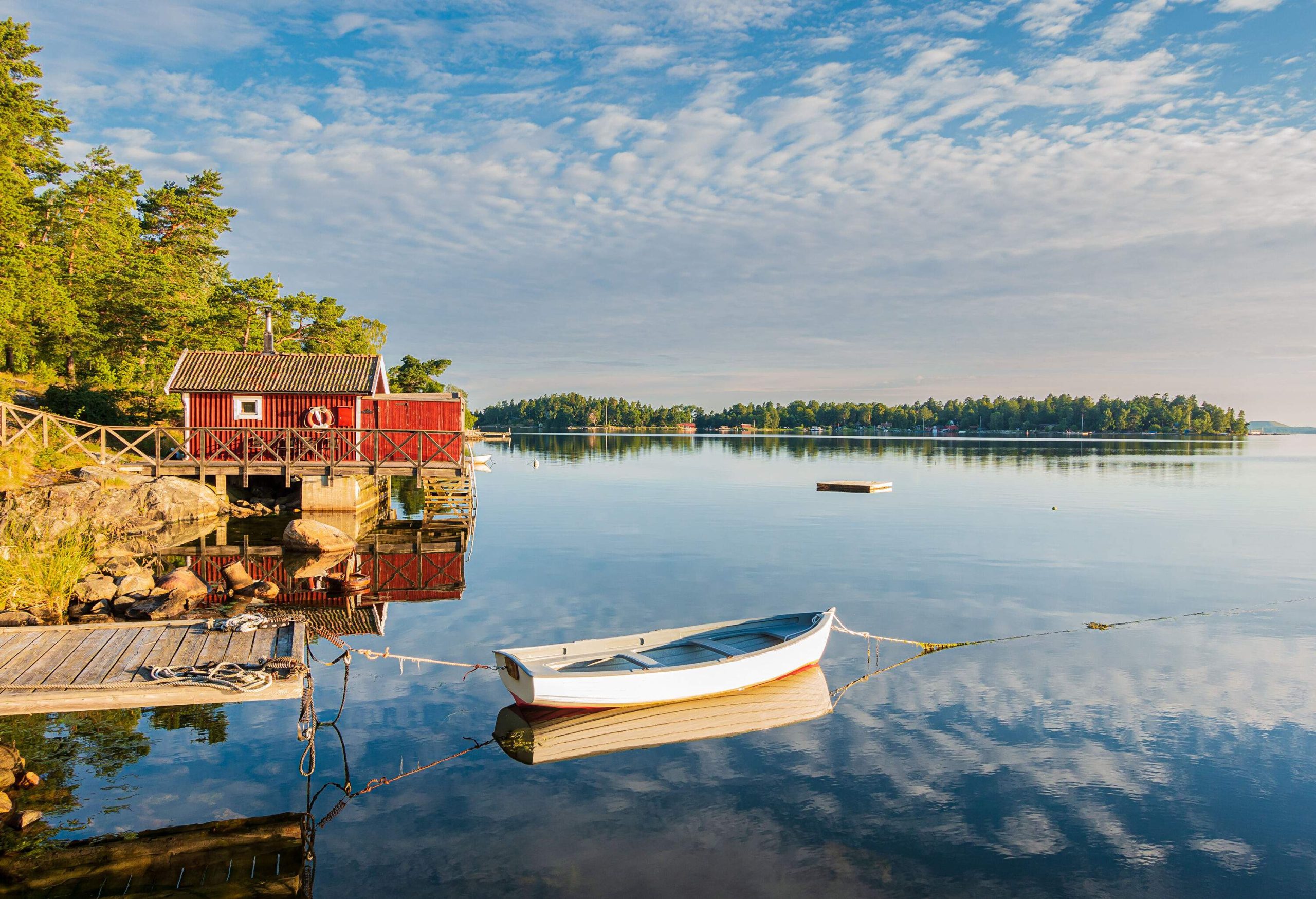 A small red house by the shore of tranquil seawater that reflects the cloudy blue sky.