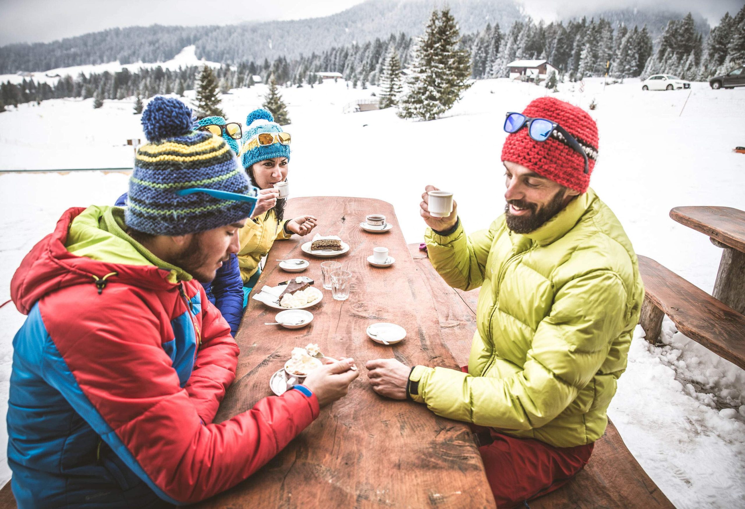 Three people dressed for the winter sitting on a wooden picnic table and enjoying good food.