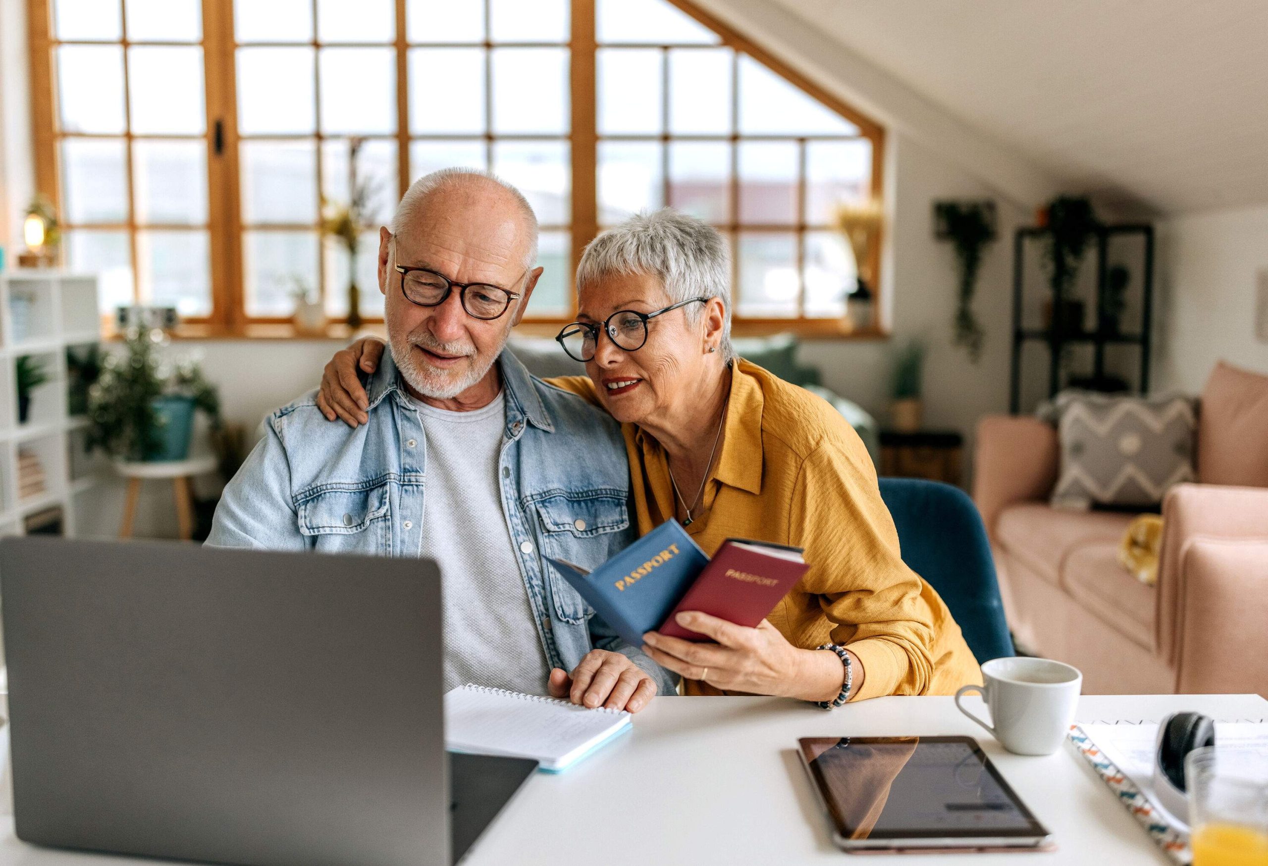 An elderly couple, the man on a laptop and the woman holding their passports, planning their trip.