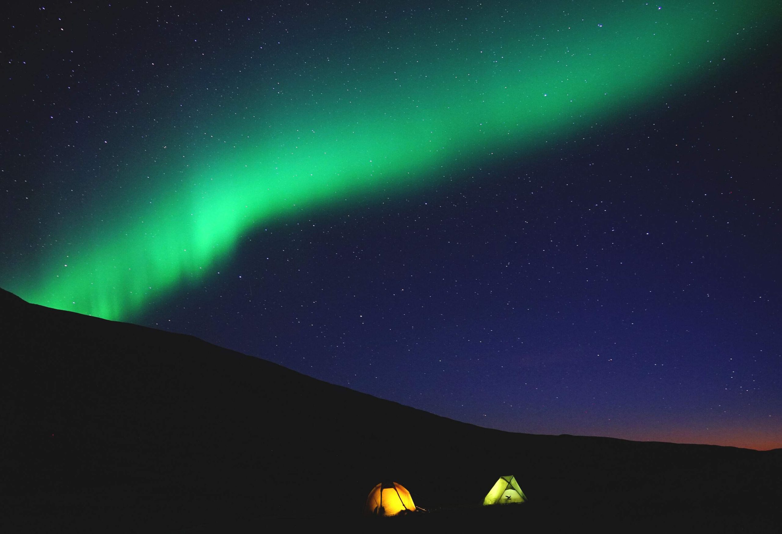 Two bright tents under a long stretch of green northern lights in the night sky.