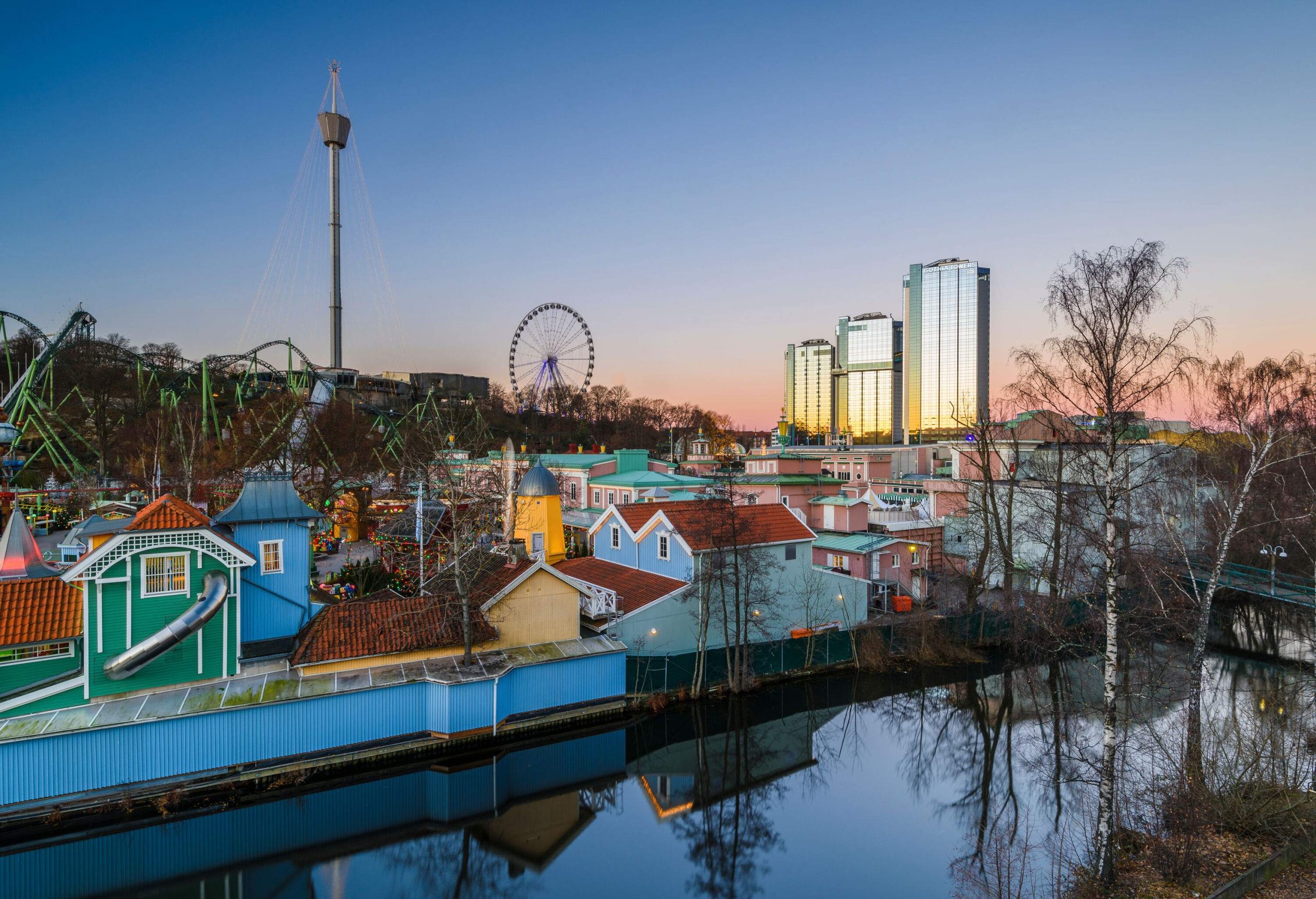 A pond along the colourful booths and structures of an amusement park with views of roller coaster rides, a Ferris wheel, drop towers, and three skyscrapers.