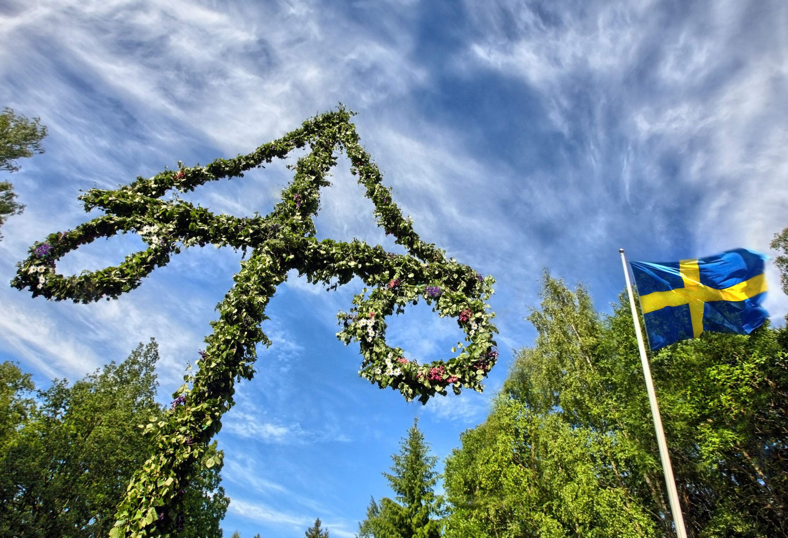 A triangular pole with hanging loops covered with leaves and flowers stands beside a Swedish flag raised on a white shaft.