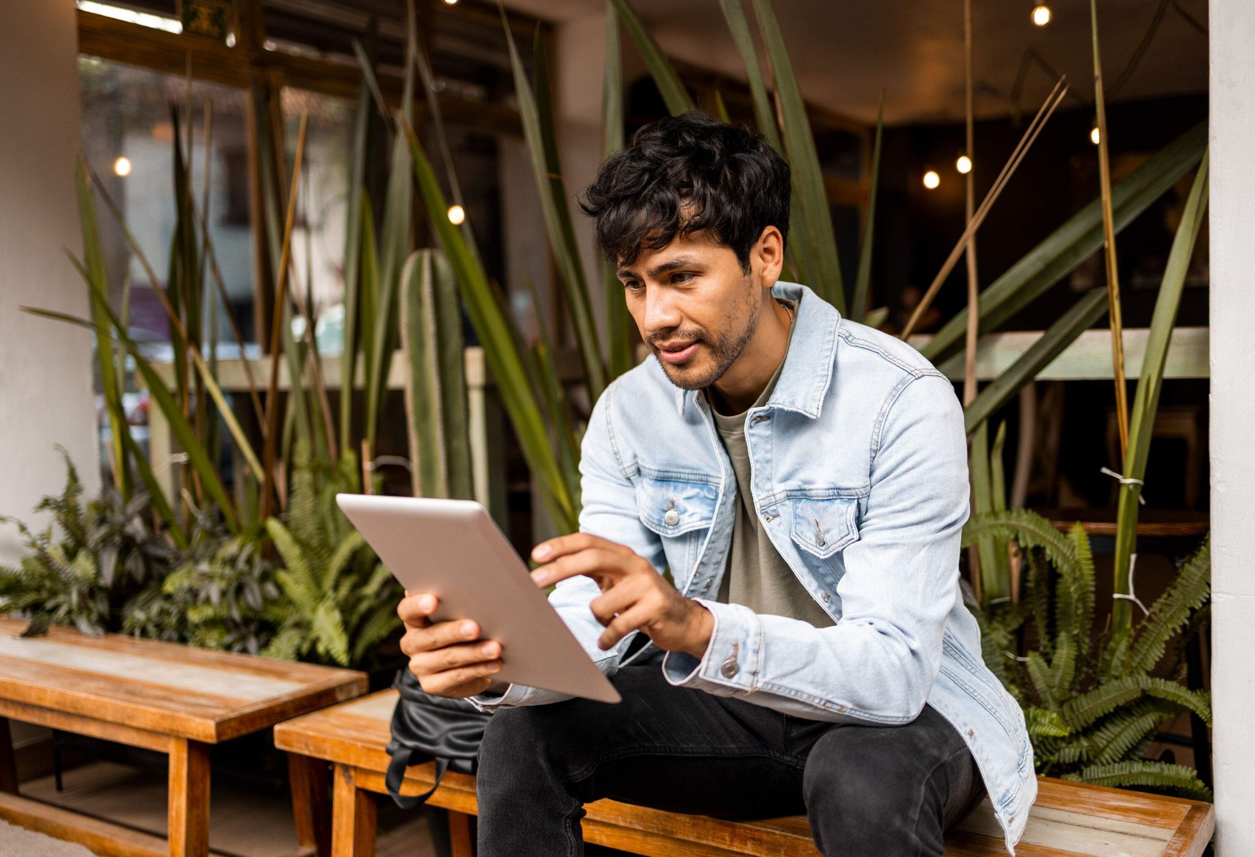 A man in a denim jacket using a tablet computer while sitting on a wooden bench.