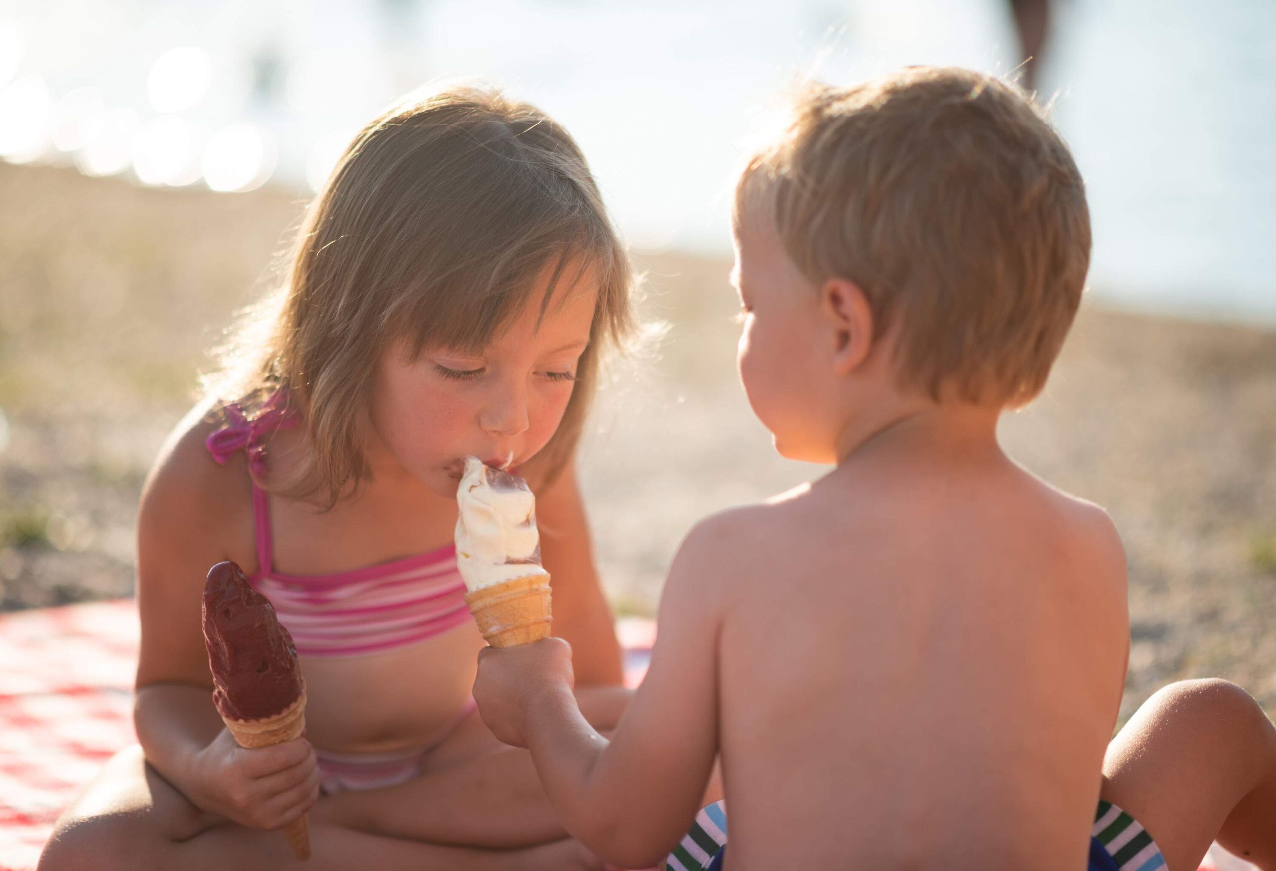 Siblings bond on the beach while enjoying cone ice cream, one playfully tasting the other's treat as they willingly share.