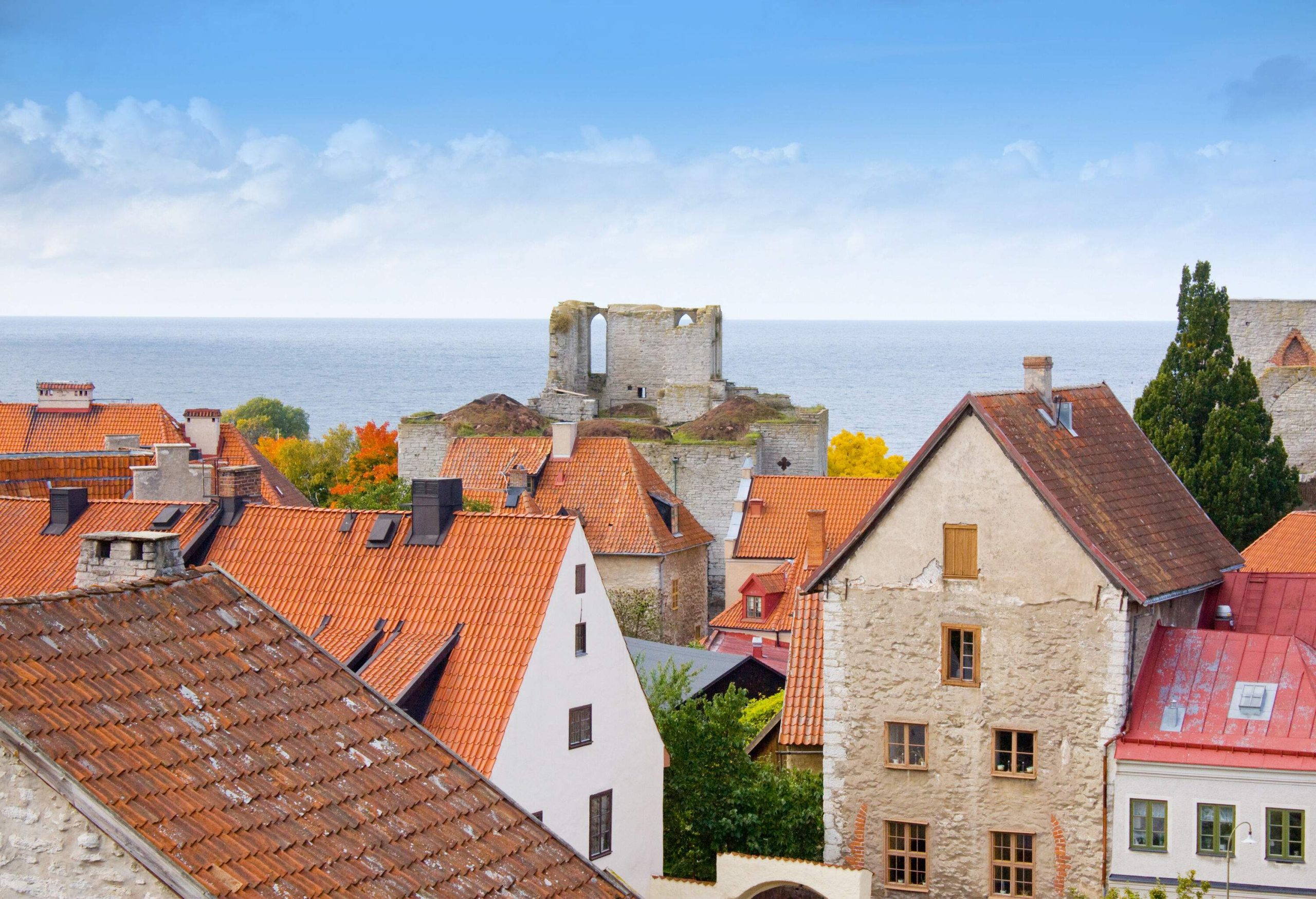 A large, beautiful light-blue sea is seen from the brick homes that are located behind the remnants of an old structure.