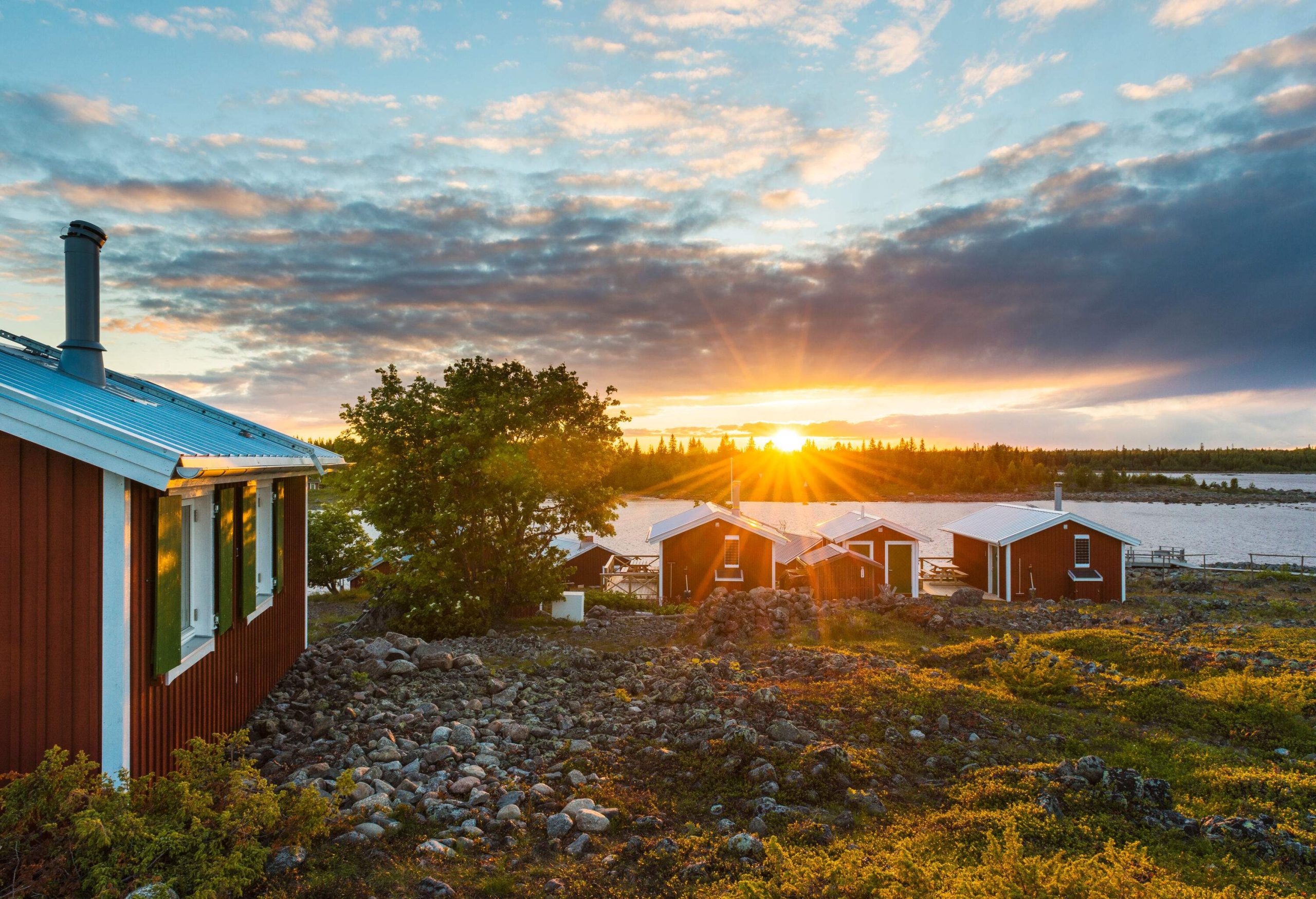 Red wooden sheds by a lake with the sun setting over the horizon.