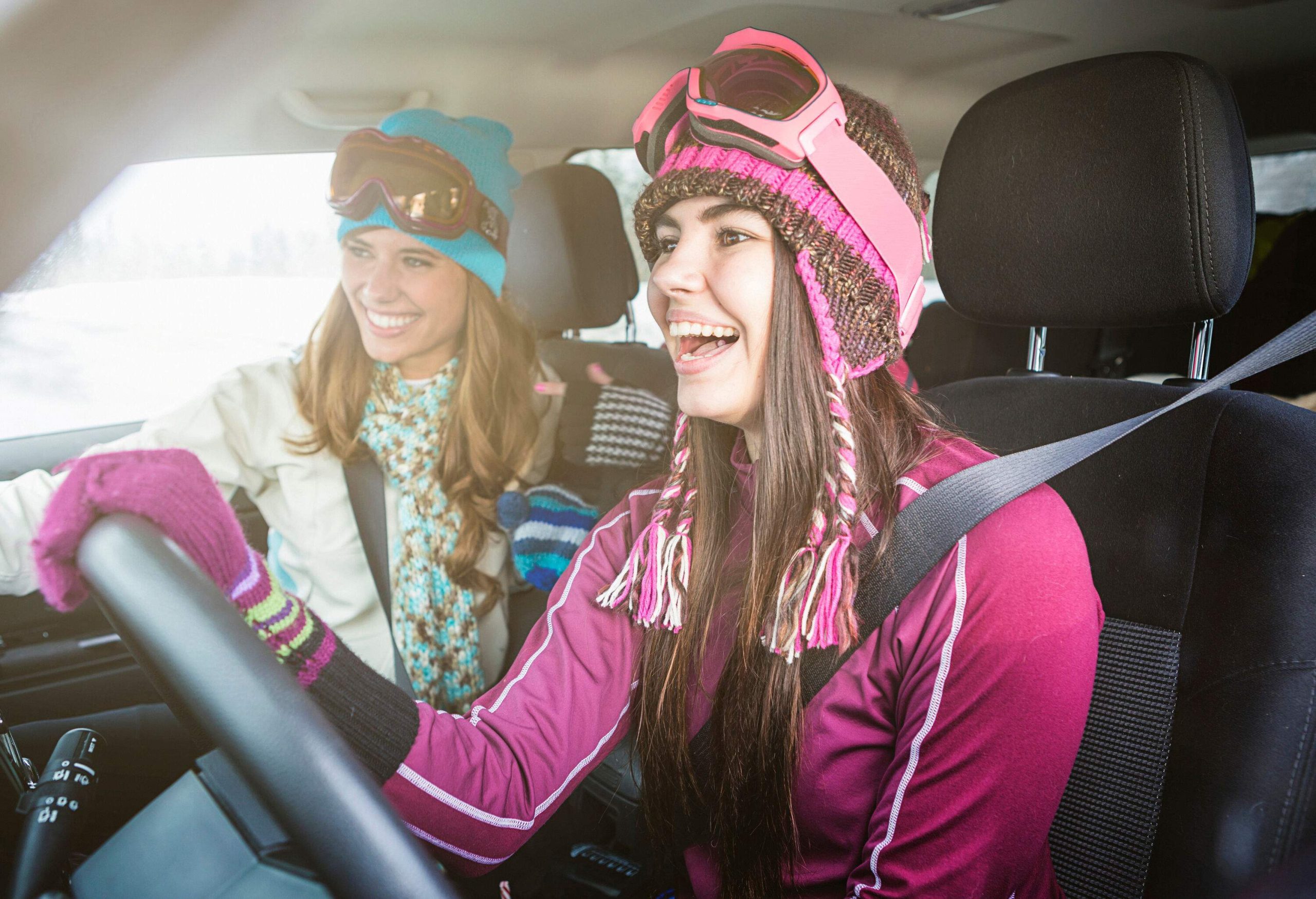Two young women dressed for skiing laughing in the front seats of a car.