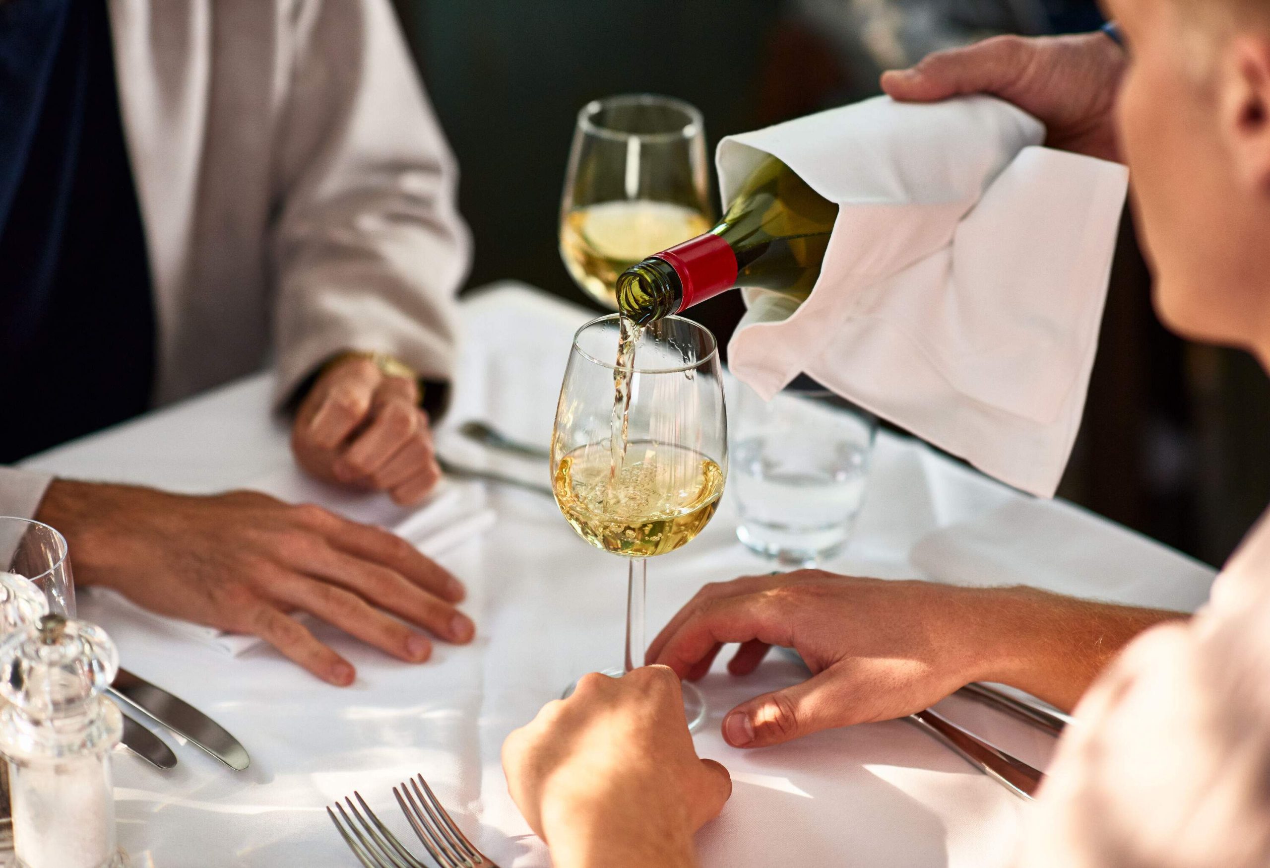 A person pours white wine for waiting customers, their hands resting on the table.