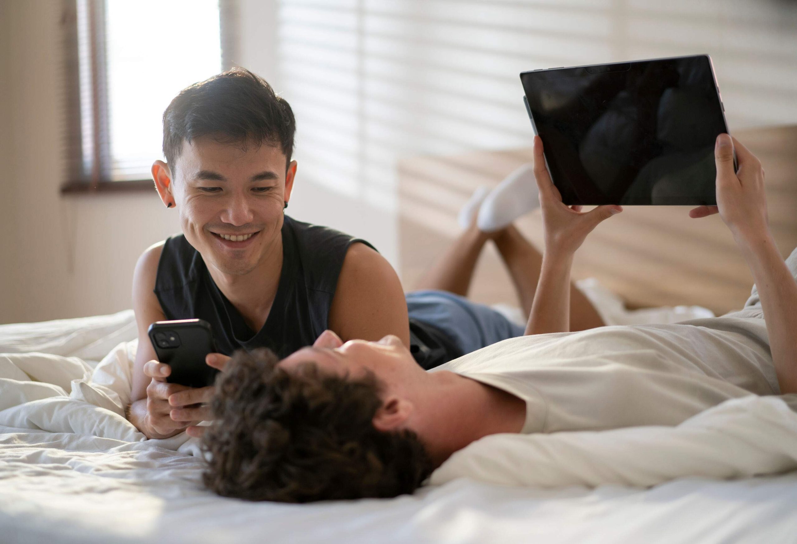 A couple lies on the bed, with one person resting on their back while the other leans against their chest, both smiling and holding a digital device.