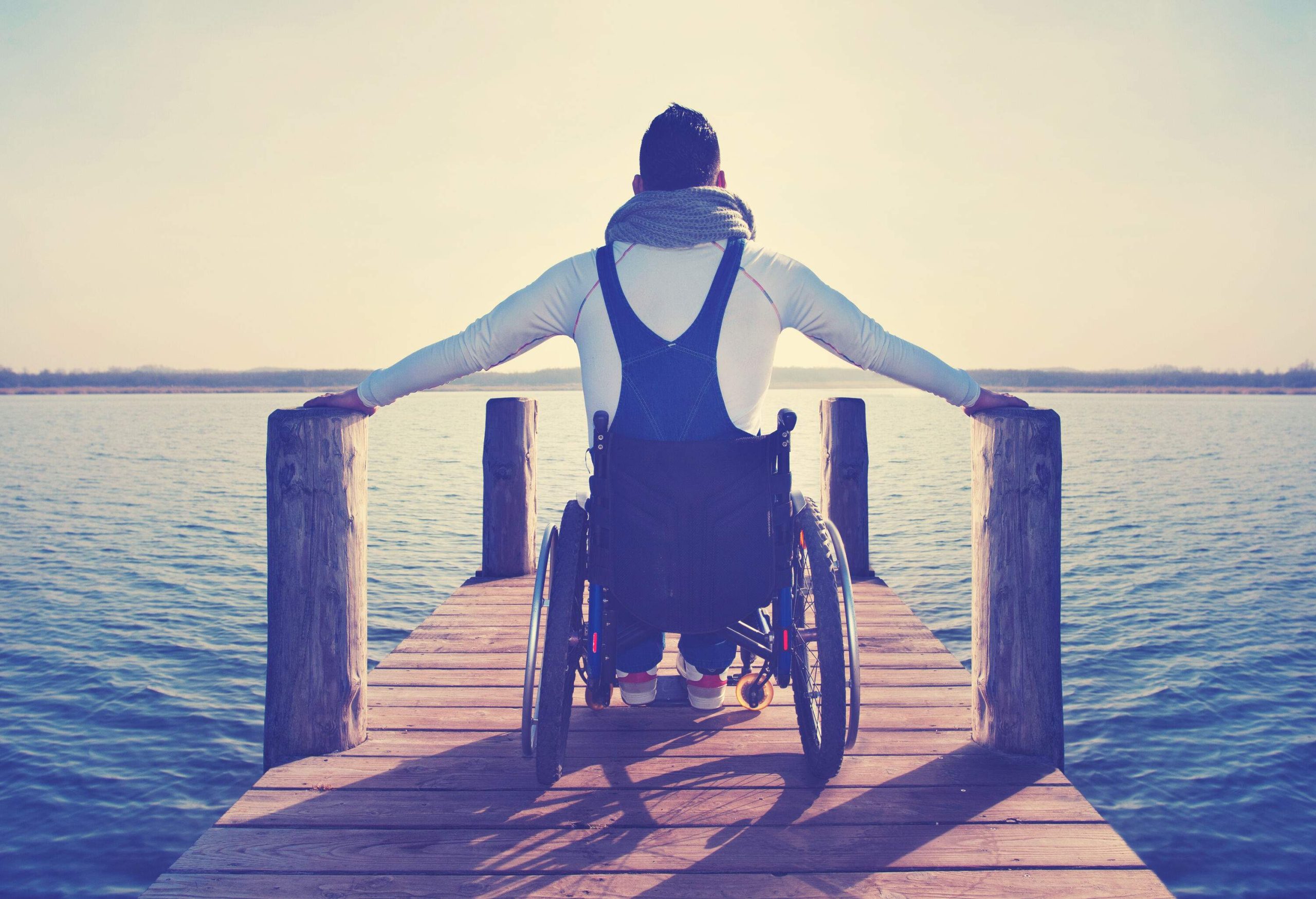A man sits in a wheelchair on a wooden pier by the lake.