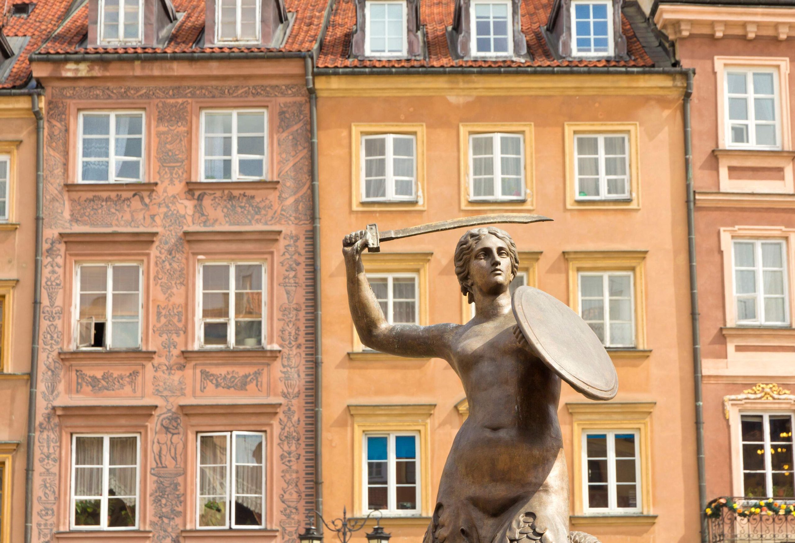 A statue of a mermaid holding a sword and a shield with colourful buildings in the background.