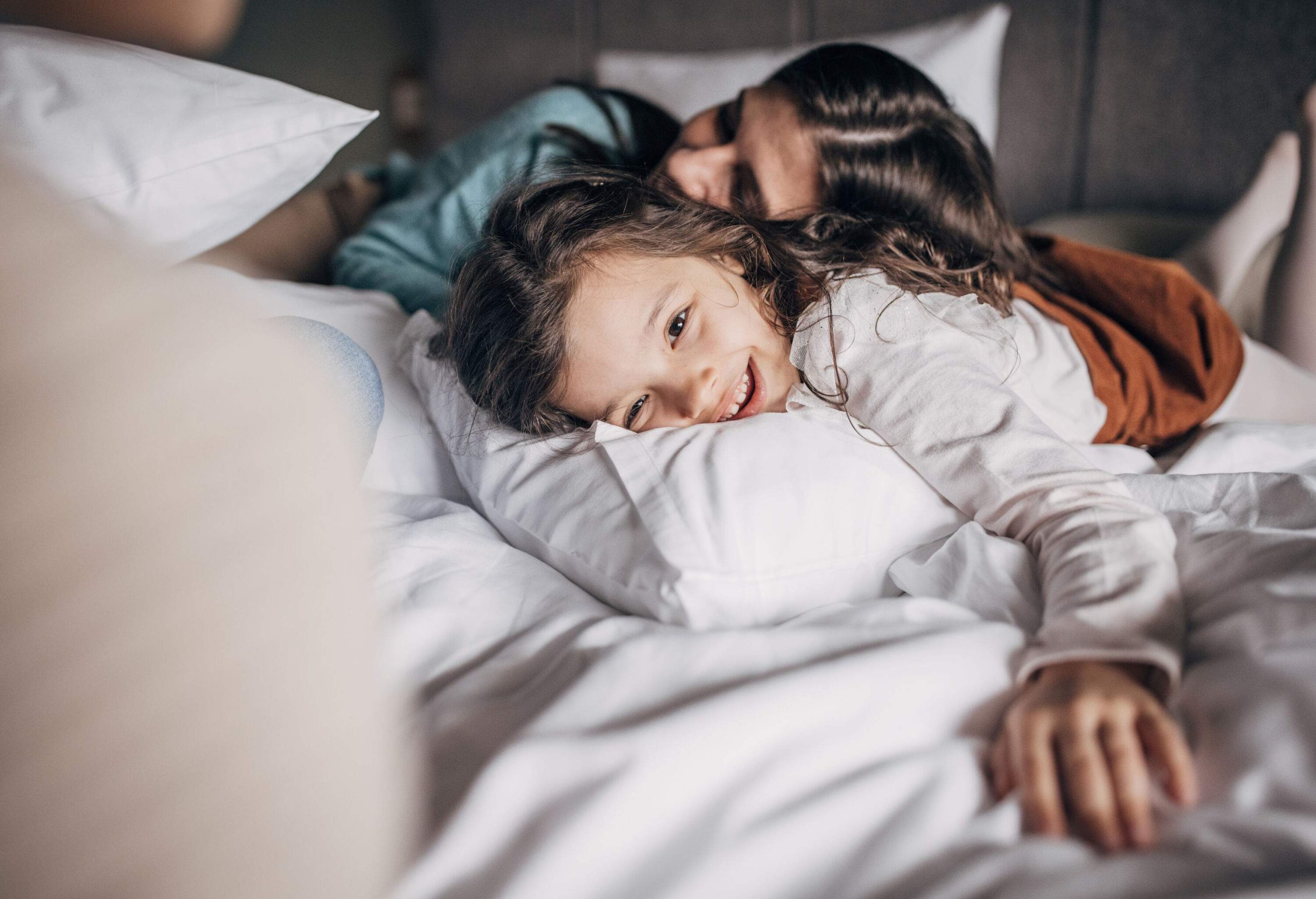 A tender moment between a mother resting her head on her daughter's back while lying on their bed.