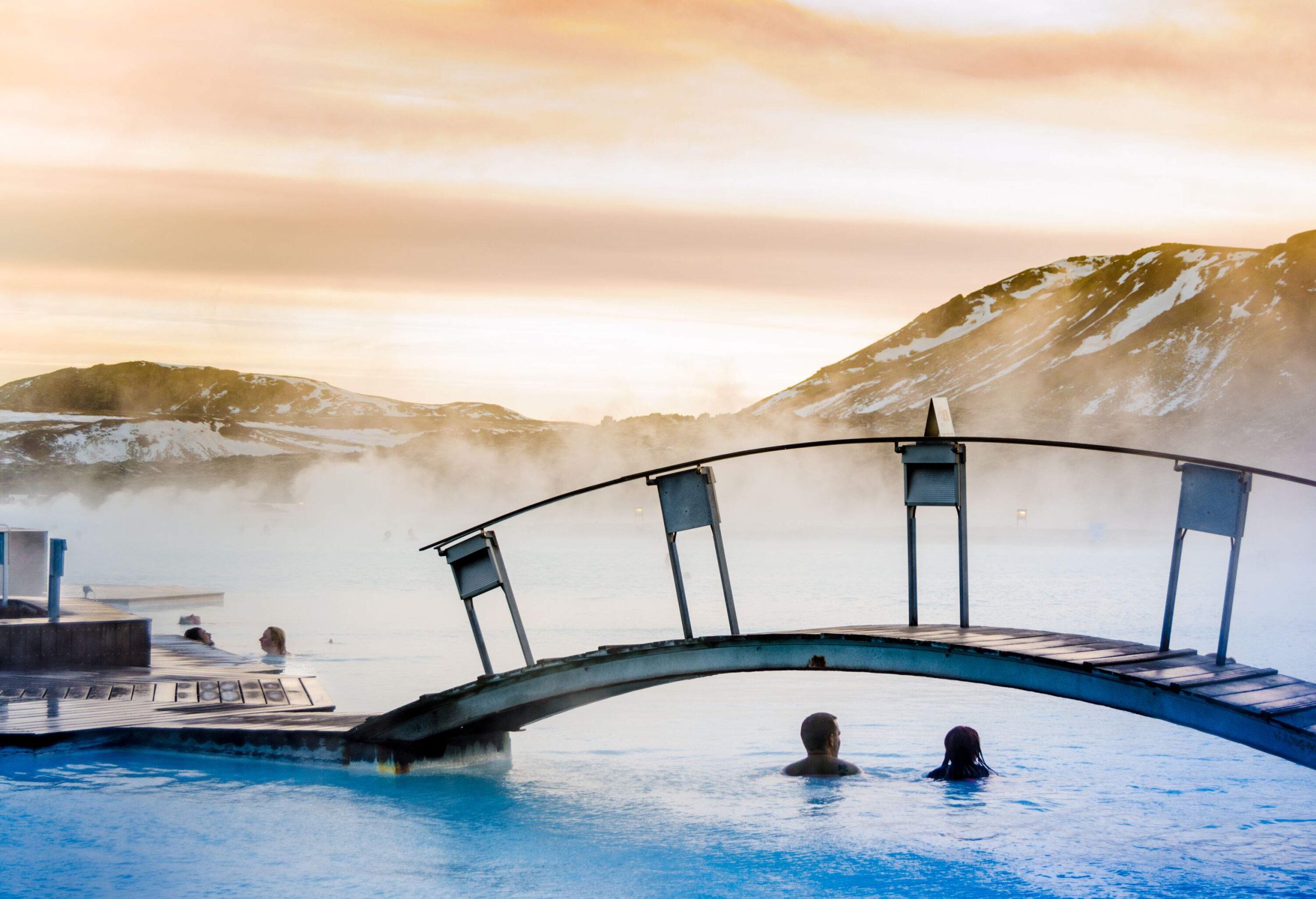 A pair of couples enjoying the blue thermal lagoon with an arch bridge surrounded by steam.