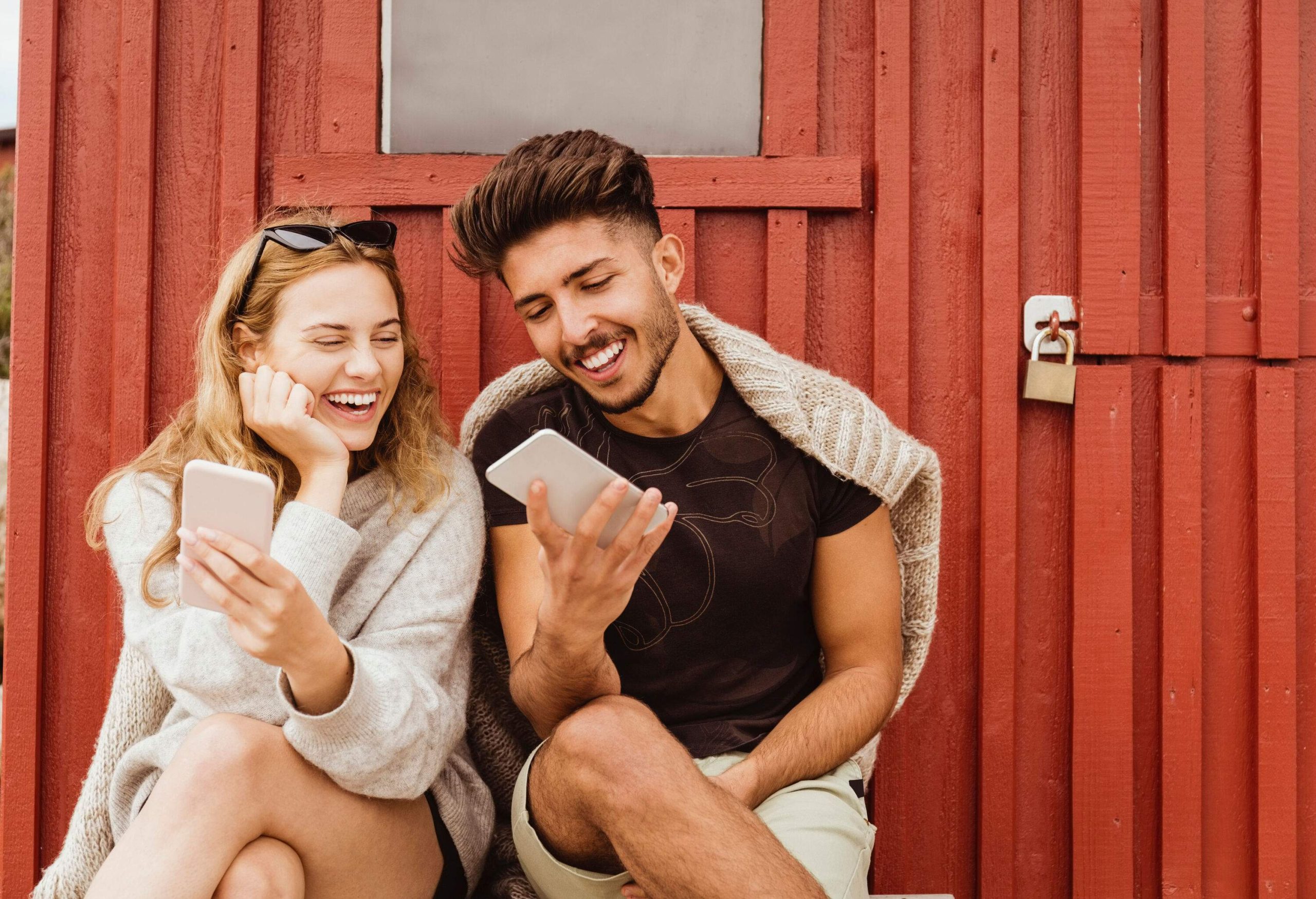 Two individuals are seated with their legs crossed and are using their mobile devices in a joyful manner.