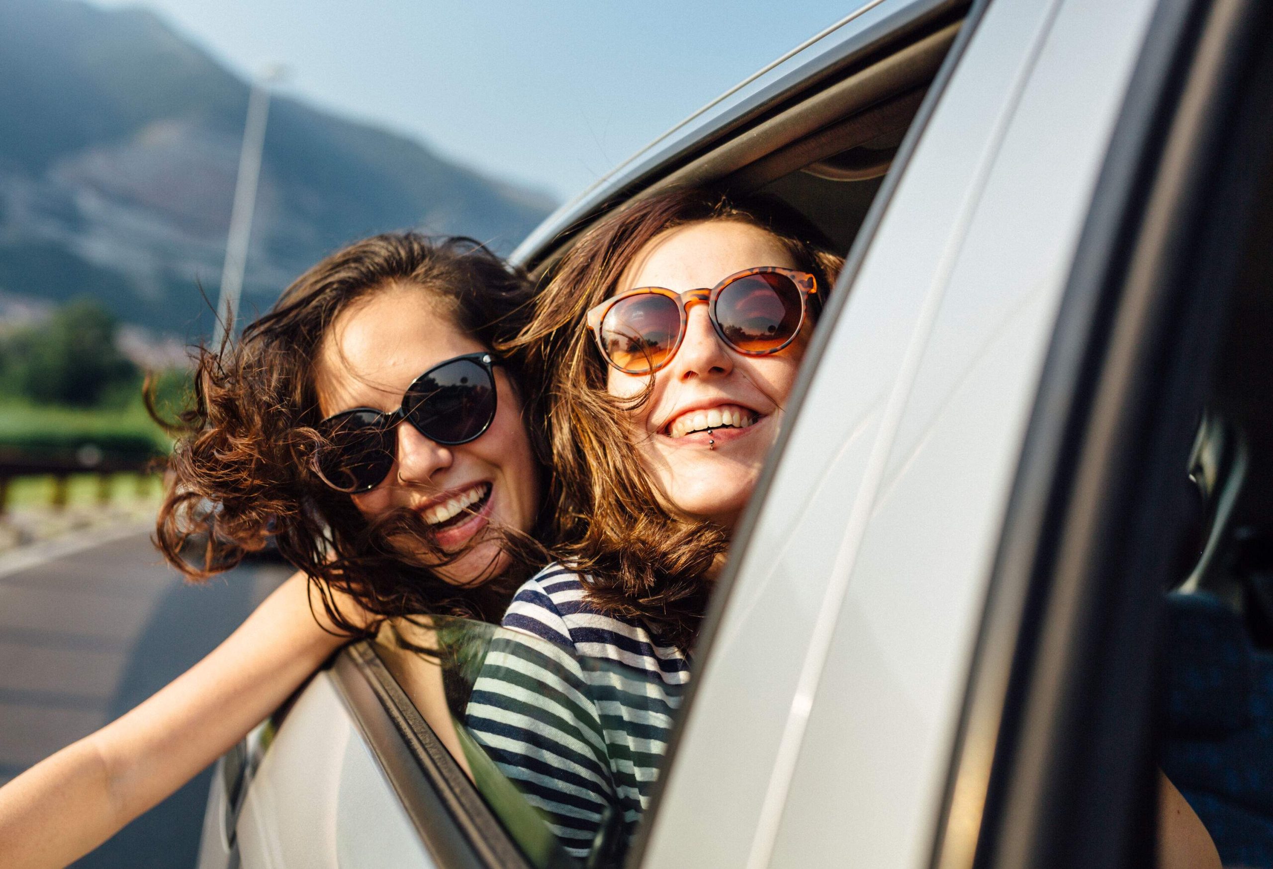 Young women friends embark on a thrilling car trip, leaning out of the car window with their heads out, basking in the refreshing breeze while sporting stylish sunglasses.
