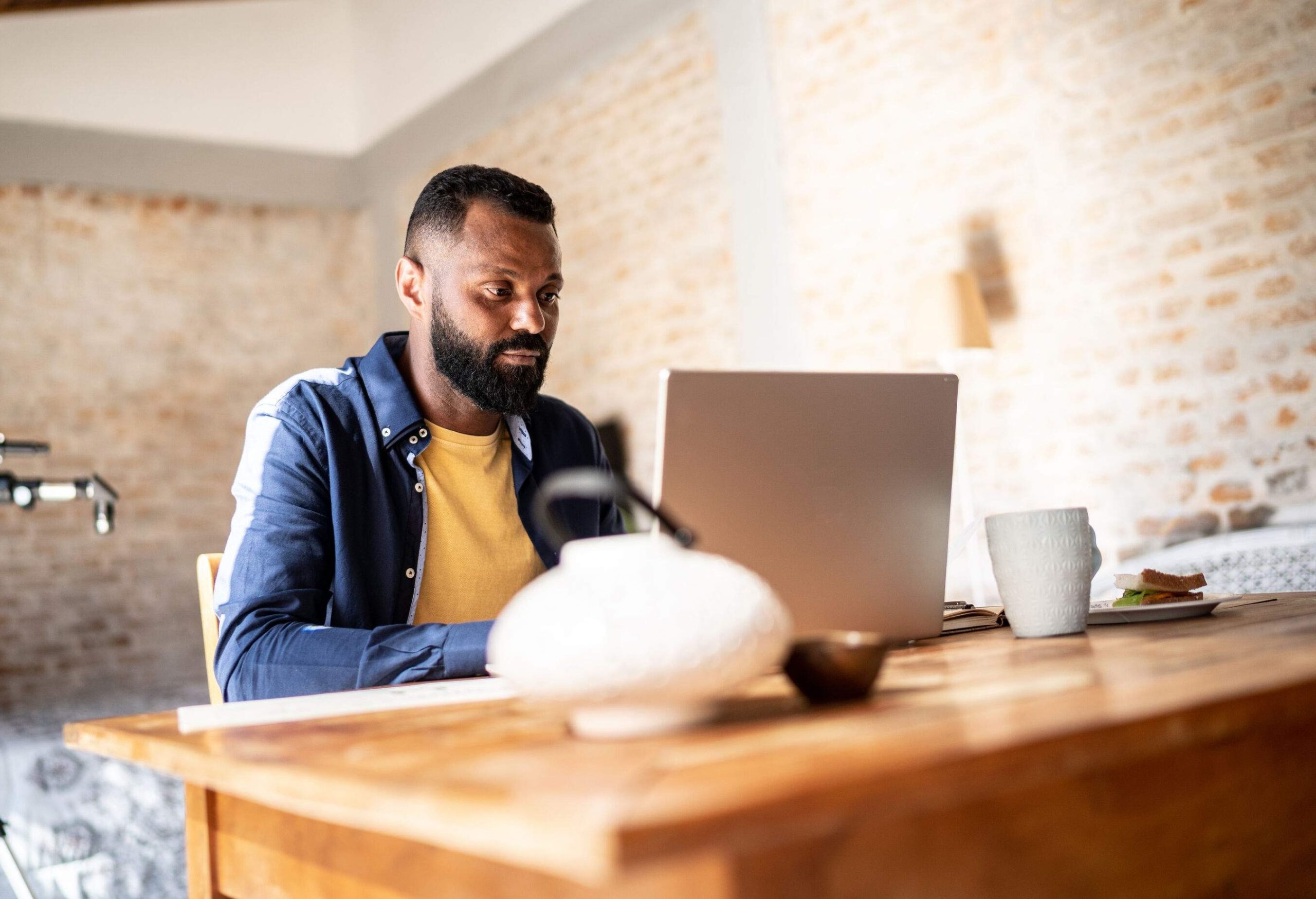 A bearded man in a blue shirt sits in front of a table while working with his laptop.