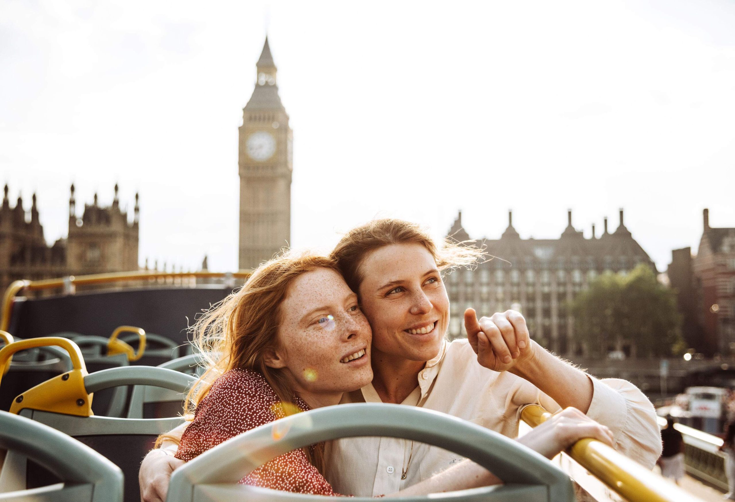 Female lovers sit and hug on an open-top bus while admiring the cityscape.