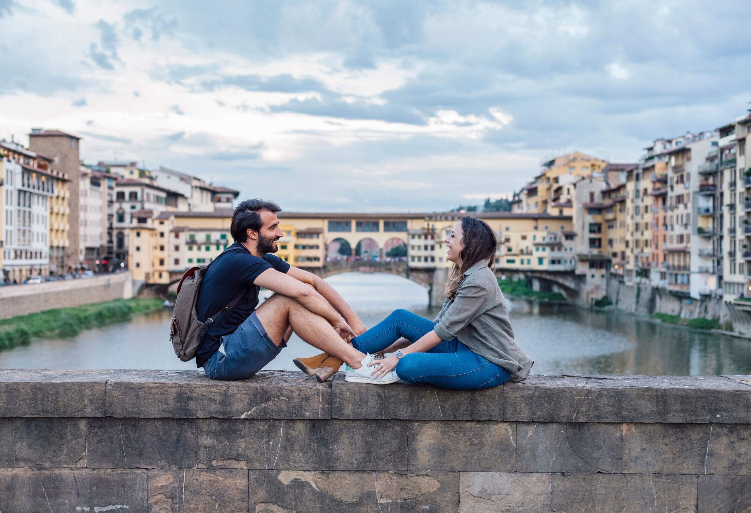 A man and a woman sitting on the wall of a bridge over a river, smiling at one other.